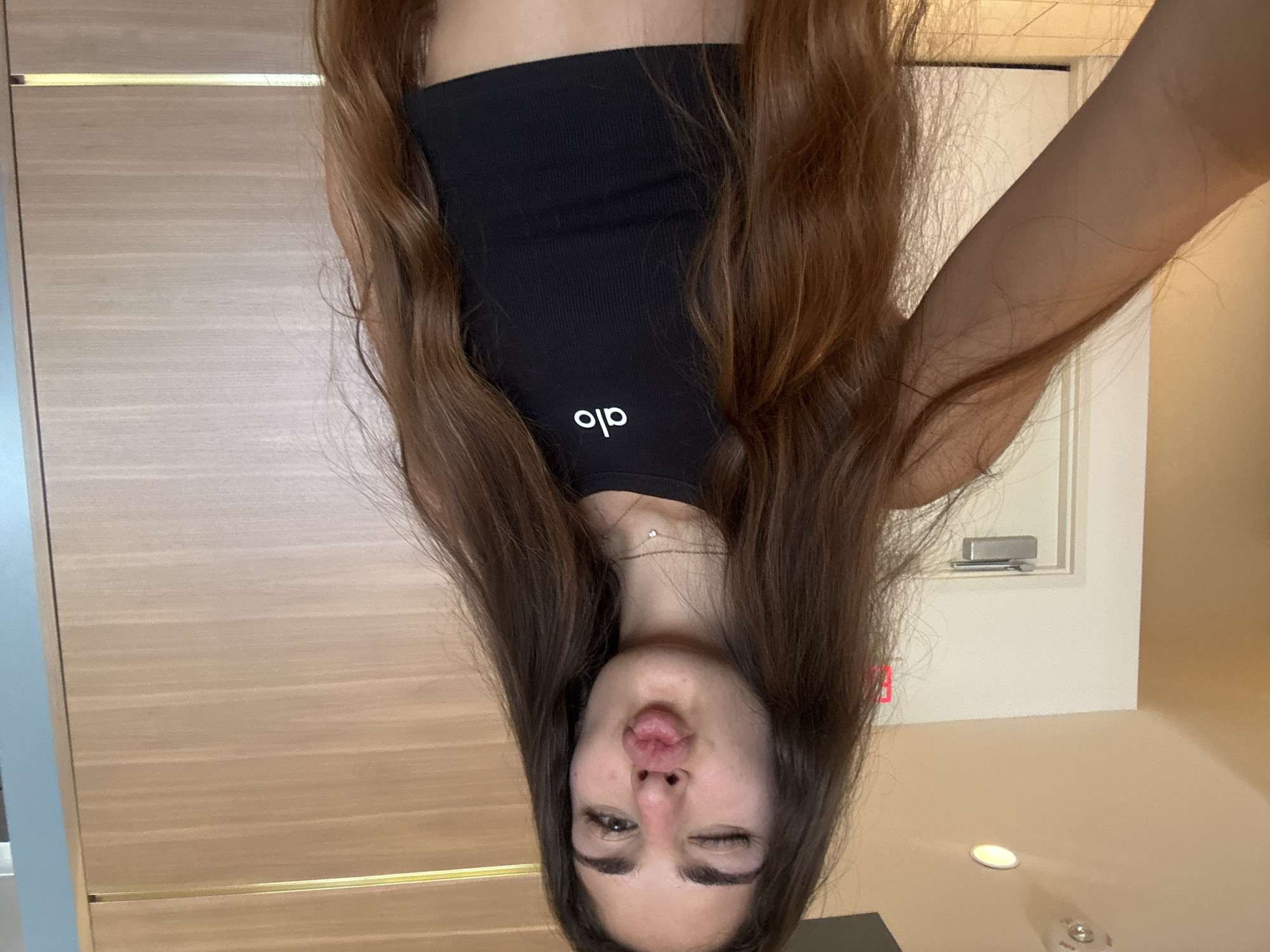 A young woman with long, brown hair is taking a selfie in a dimly lit room. She is wearing a black ribbed tank top with the white logo "alo" visible on the front. Her hair is falling over her shoulders, partially obscuring her face, and she is making a kissy face while winking. A thin, delicate necklace with a small charm is visible around her neck. The background shows wooden paneling on the left and a cream-colored wall on the right with a framed picture on it. The image appears to be taken indoors, likely in a residential or commercial setting. The lighting suggests it could be evening or an indoor environment without bright natural light. There is no indication of the weather or specific time of day beyond the interior lighting. The overall mood seems playful and casual.