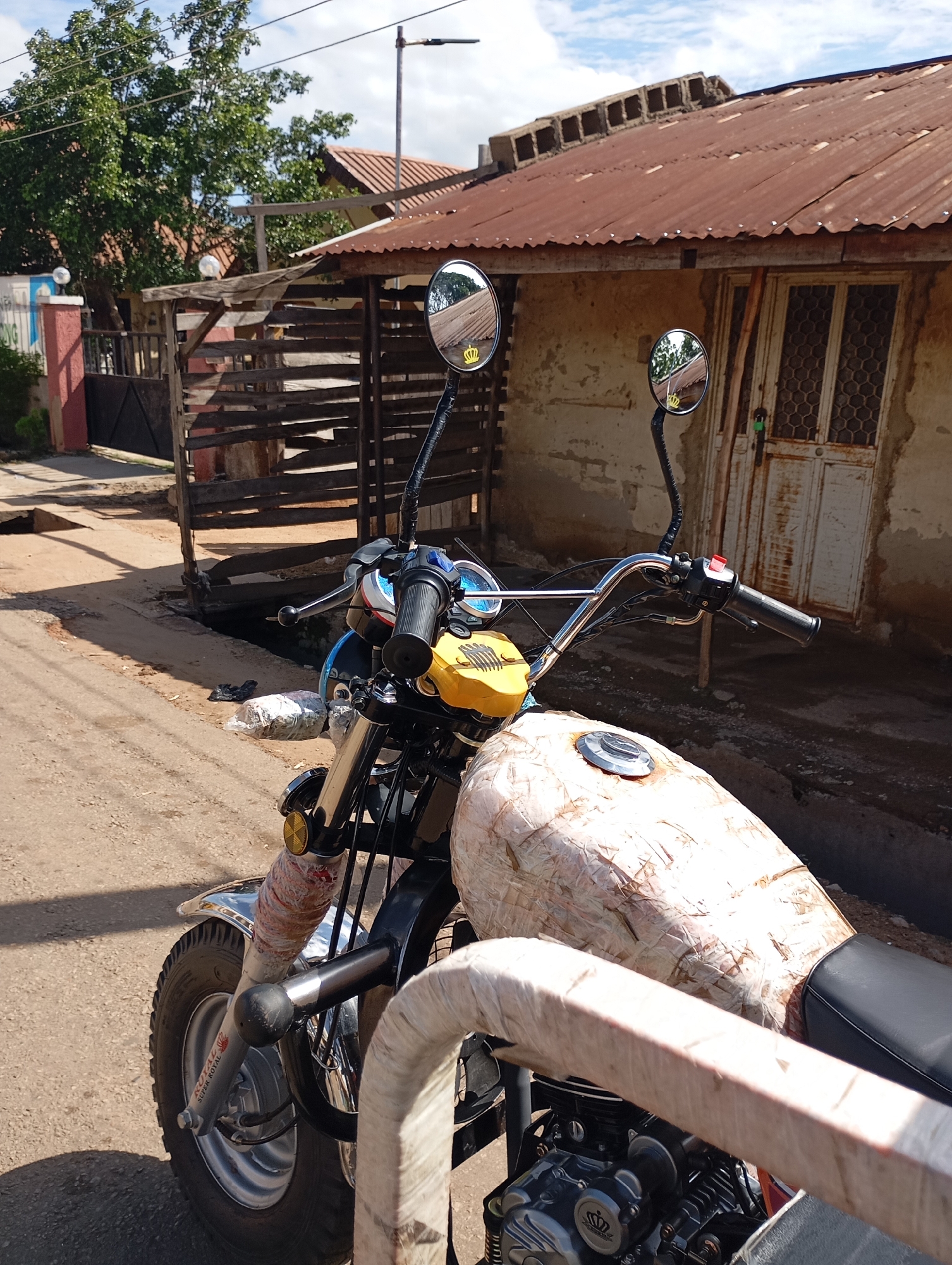 A motorcycle is parked on a dirt road in front of a weathered, corrugated metal-roofed building. The motorcycle's fuel tank is wrapped in a light-colored material, possibly for protection or decoration. The handlebars are visible, with mirrors reflecting the surroundings. The building appears to be made of rough plaster and has a wooden door with a lattice pattern. A wooden structure, possibly a fence or shelter, is situated to the left of the building. Vegetation, including a large tree, is visible in the background. The sky is partly cloudy. This scene is located in Shendam, Nigeria.