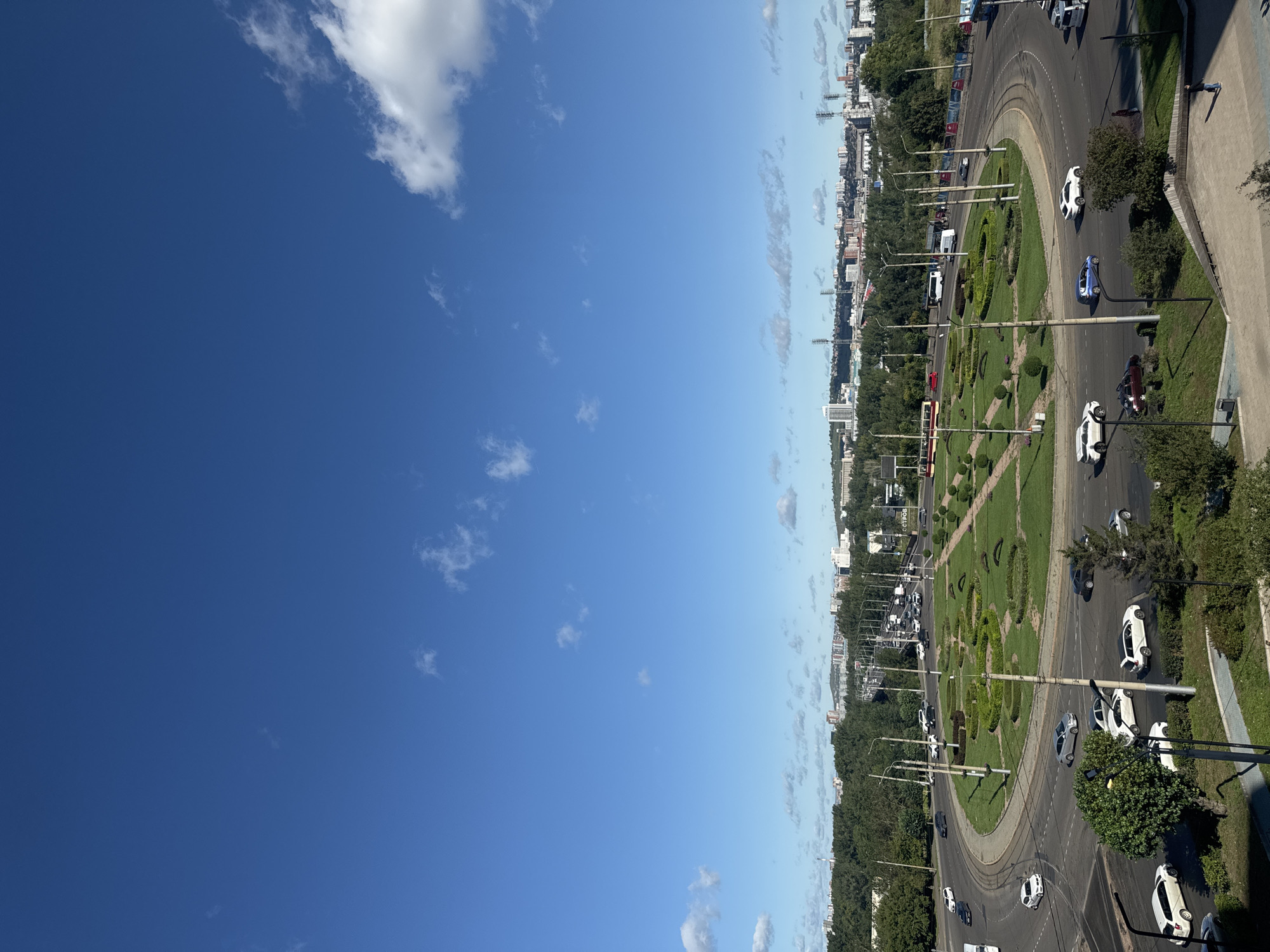 The image shows a wide-angle view of a large, circular intersection with a landscaped median in the center, located in the city of Krasnoyarsk, Russia. The sky is a clear, bright blue with scattered white clouds. The intersection is surrounded by green trees and a cityscape of buildings in the background, with several tall transmission towers visible.
Numerous cars and a tram are actively navigating the roundabout and surrounding roads. The landscaping in the center of the roundabout features manicured green grass arranged in decorative patterns and small trees. Light poles are strategically placed throughout the intersection and along the roads.
In the foreground, on the right side, there are stairs and a paved area with a few pedestrians. The overall scene depicts a busy urban environment on a sunny day.
