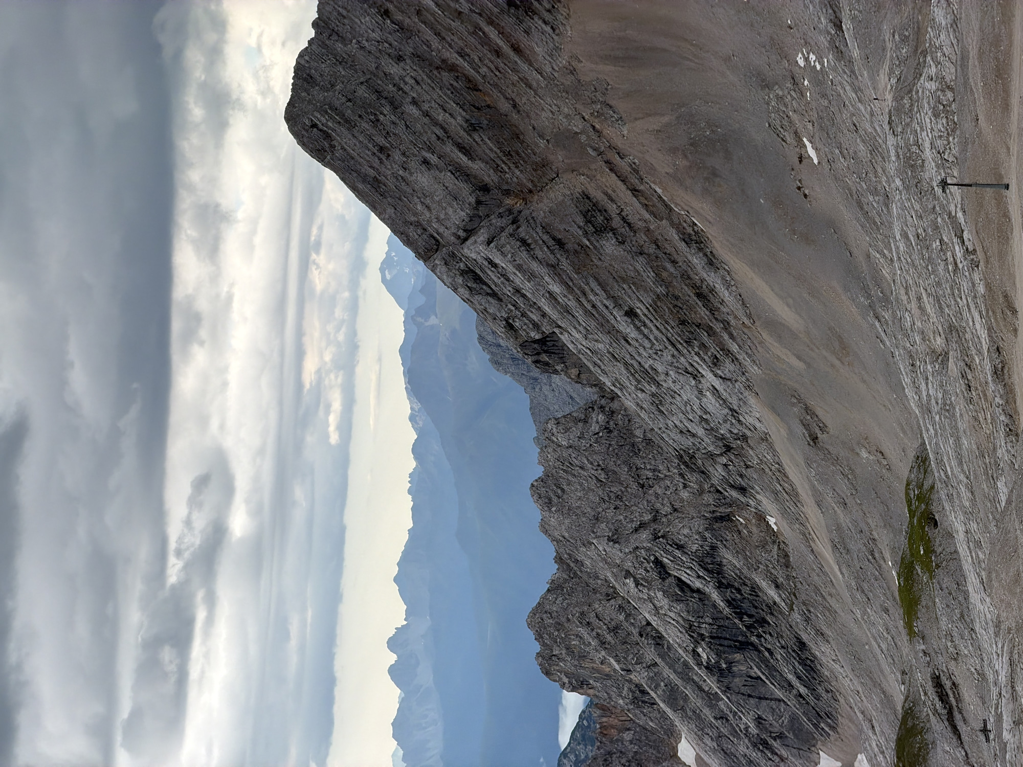 The image depicts a mountainous landscape, likely the Zugspitze, near Garmisch-Partenkirchen, Germany, under a cloudy sky. The foreground is dominated by a rugged, rocky slope covered in scree and patches of sparse vegetation. Jagged mountain ridges with distinct striations stretch across the midground. In the distance, layers of mountain ranges fade into a hazy blue. A single, dark ski lift pylon stands in the lower right quadrant of the image, and a small, silhouetted figure can be seen walking on the slope further up and to the left of the pylon. The overall impression is one of a vast, untamed alpine environment.