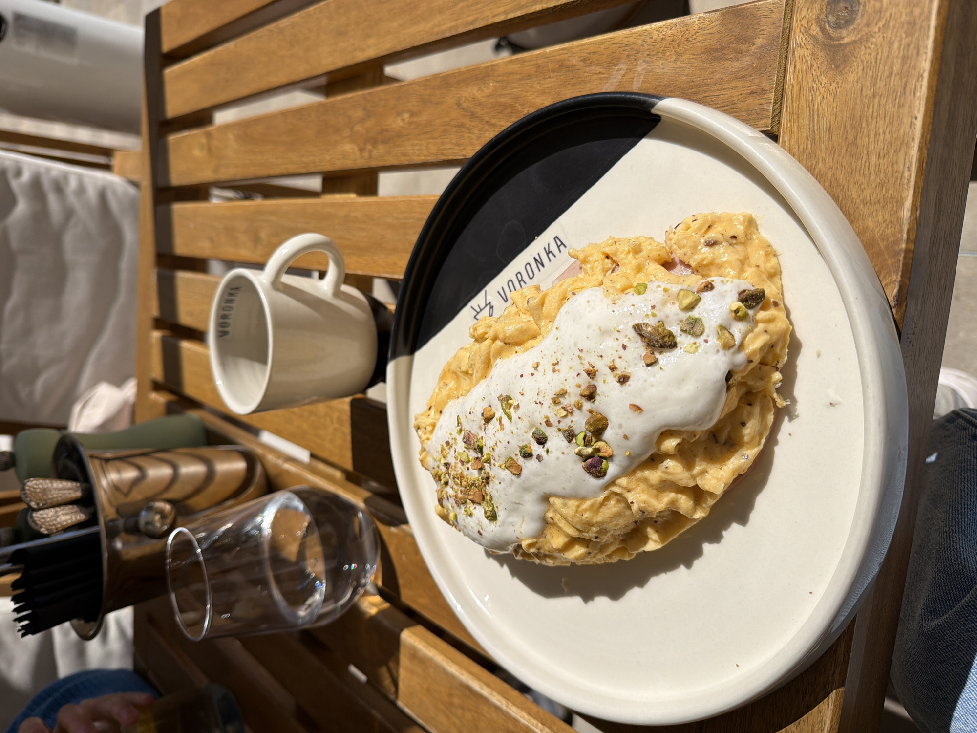 The image captures an outdoor dining scene in Boreti, Montenegro, featuring a plate of food on a wooden slatted surface under bright sunlight.
The main subject is a dish presented on a round, white ceramic plate with a distinctive black and white geometric design on its rim. The food appears to be a fluffy omelet or scrambled eggs, topped with a generous dollop of white, creamy sauce and garnished with crushed green pistachios. The word "VORONKA" is visibly inscribed on the plate's rim.
To the left of the plate, a white ceramic mug, also bearing the "VORONKA" logo, is placed on the wooden surface. Below it, a clear drinking glass is visible. Further left and slightly out of focus, there's a rustic wooden holder containing various utensils, including what appear to be metal straws or stirring rods with decorative ends.
The setting is an outdoor space, likely a balcony or a cafe patio, characterized by the natural wooden slatted furniture. Bright, clear daylight is evident, casting distinct shadows and suggesting a sunny morning or early afternoon. A partial view of a person's leg, dressed in blue jeans, is visible in the bottom right corner, implying a diner is seated at the table. The overall scene conveys a relaxed outdoor meal.