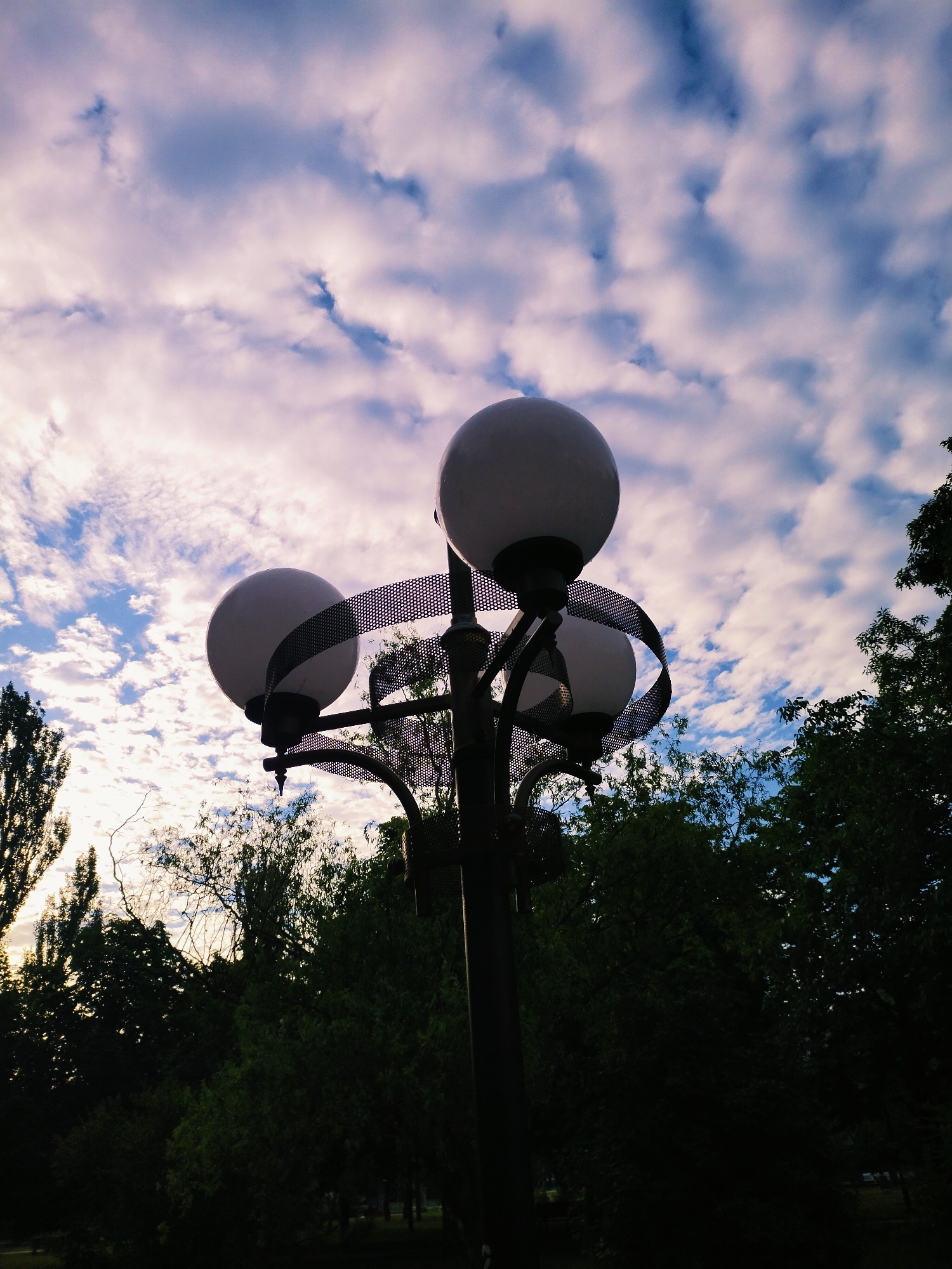 A street lamp with three spherical globes stands silhouetted against a cloudy sky in Horishni Plavni, Ukraine. The lamp is dark, with a metal grid wrapped around its central pole. The globes are illuminated, casting a soft light. Dark green trees are visible in the background, with some lighter trees on the left side of the image, suggesting it is either dawn or dusk. The sky is a mix of blue and white clouds, with hints of purple and pink. The ground is not visible in the image.