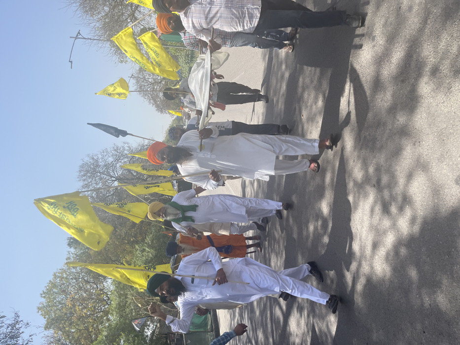 The image depicts a group of men walking down a street in Sahibzada Ajit Singh Nagar, India. The men are carrying a white cloth stretcher. They are all wearing turbans and traditional clothing. There are yellow and green flags with Punjabi writing on them planted along the street. The men are walking in a procession. The image is taken during the daytime, under clear skies. The street appears to be paved with asphalt and has some shadow cast by the trees.