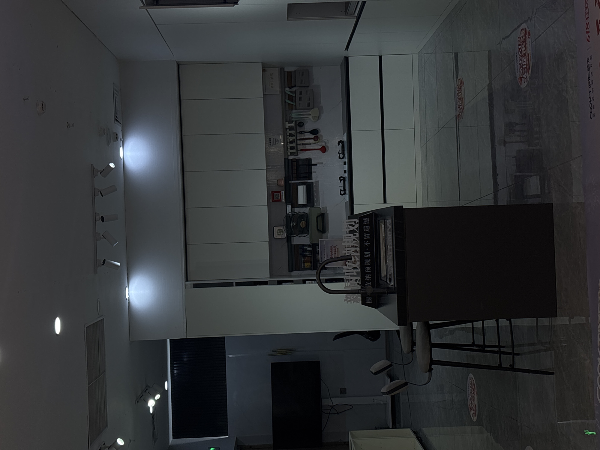 Here is a detailed description of the image:
**Scene Overview:** The image shows a kitchen exhibit or showroom, likely located in a retail or commercial space. The photograph appears to be taken from an eye-level perspective, slightly off-center.
**Subjects and Objects:**
* **Kitchen Cabinetry:** The prominent feature is a set of modern kitchen cabinets with white panels. These cabinets occupy a significant portion of the backdrop.
* **Display Elements:** The cabinets have various kitchen utensils, appliances, and cookware displayed as part of the exhibit.
* **Countertop and Sink:** A dark-colored countertop with a sink and faucet is visible.
* **Display Stand:** A dark-colored display stand or counter with shelving is situated in the foreground.
* **Lighting:** There is a strip of lights affixed to the wall.
* **Television:** A television is affixed to the wall.
**Environment:**
* **Flooring:** The floor appears to be tiled, with decorative circular designs.
* **Walls:** The walls are white.
**Location Information (implied):** The location is specified as the city of Liangjiang, China. However, there is no information about the city of Liangjiang from the context of the image.
**In Summary:** The image depicts a kitchen display, most likely in a showroom or retail setting, that features cabinets, a sink, and decorative elements to showcase kitchen design and products.
