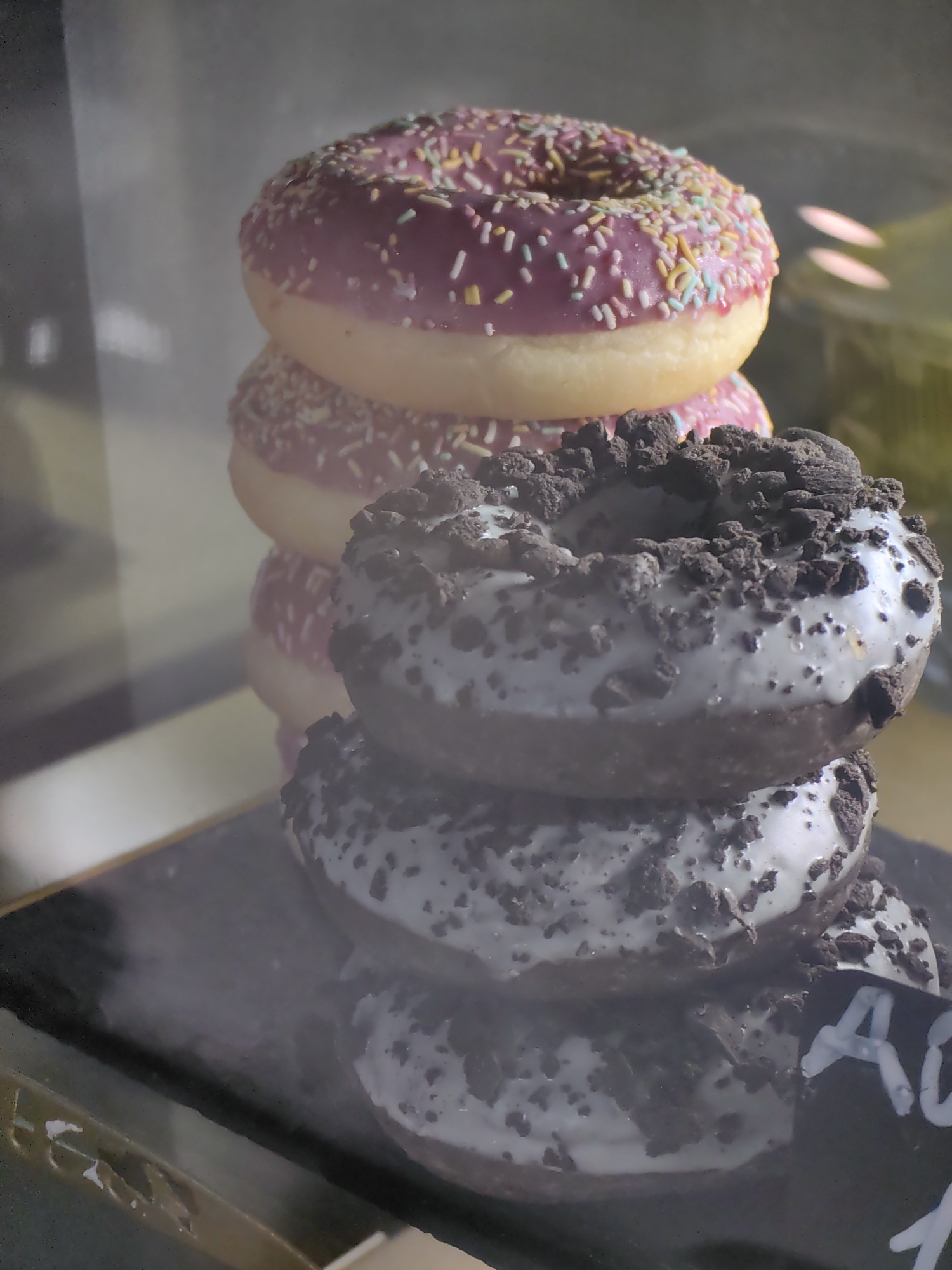 The image shows a close-up, stacked display of donuts. The donuts are arranged vertically, with the top three appearing to be a variety of pink-frosted donuts with colorful sprinkles, and the bottom two being dark chocolate donuts with white icing and crumbled cookie toppings. The lighting is dim, suggesting an indoor setting, possibly a bakery or cafe display case, given the reflective surfaces and slightly hazy quality of the image. The focus is sharp on the donuts, highlighting their textures and toppings. To the right, partially obscured, is a dark surface with some white writing, though the text is mostly illegible except for a few letters that appear to be "AO". No people are visible in the frame, and the overall mood is suggestive of a sweet treat display. The time of day and weather are not discernible.