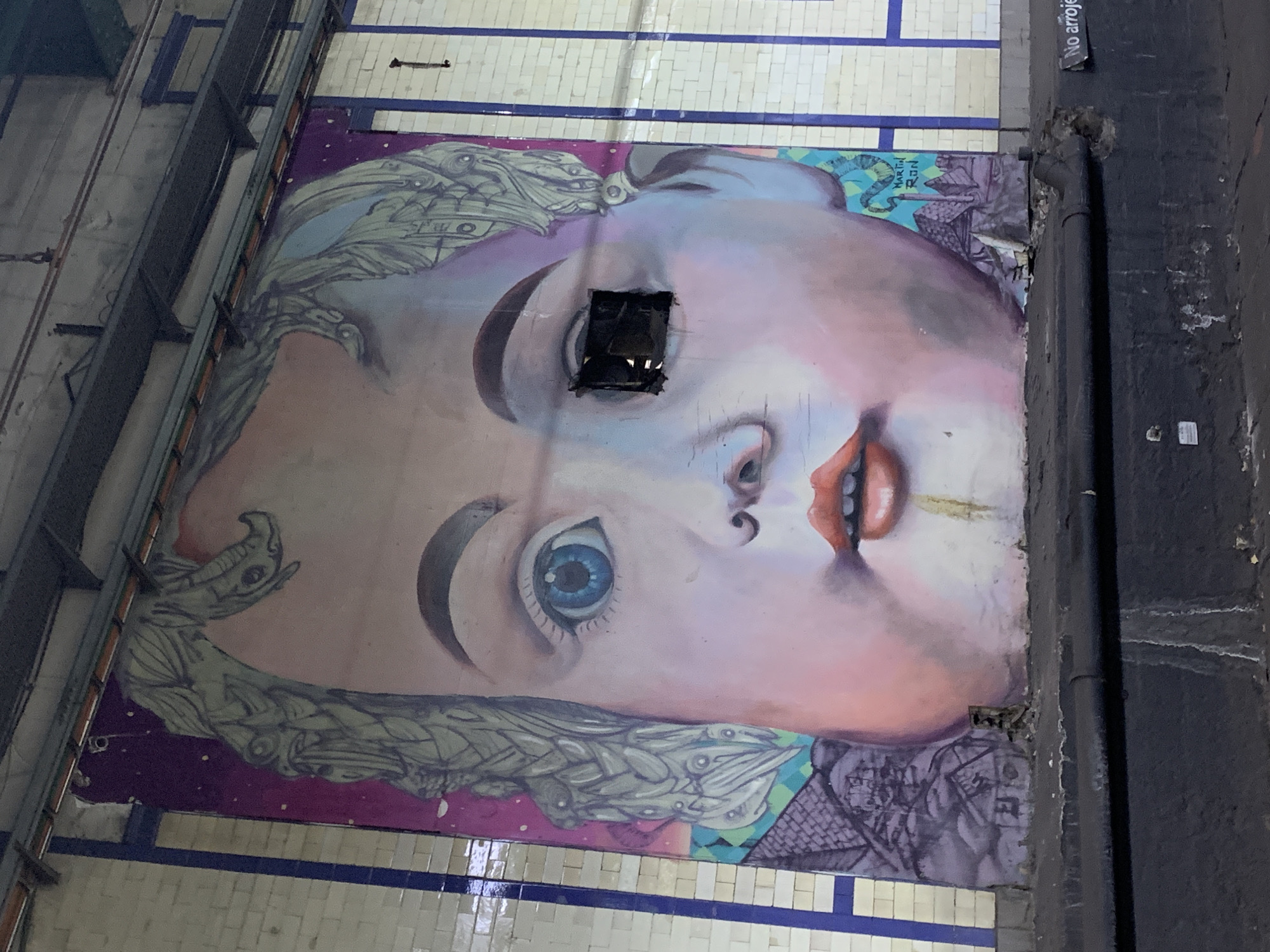 The image shows a mural of a large face on a wall, likely in an underground or industrial setting due to the visible pipes, beams, and tile work. The face is pale with detailed features like blue eyes and red lips. A square opening is cut out near one of the eyes. The mural is surrounded by structural elements and a tiled wall with a blue stripe. The mural is in Buenos Aires, Argentina.