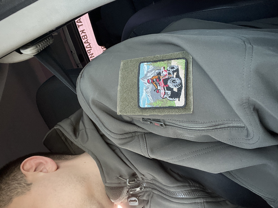 A male individual is observed seated within a vehicle's interior, visible from the right profile of his head and upper torso. He wears an olive-green tactical softshell jacket featuring multiple zippered pockets, including a chest pocket on the wearer's left with a small red 'H' logo (consistent with Helikon-Tex branding), and a zippered bicep pocket. A square, embroidered patch is attached to the right bicep's hook-and-loop panel. The patch depicts a red all-terrain vehicle (ATV) with a rider wearing an orange helmet and shirt on a dirt path, set against a background of green trees, blue sky, and snow-capped mountains. The vehicle's interior includes a dark seat, a light-colored pillar on the right, and a black seatbelt. Through the vehicle's window, reversed Cyrillic text resembling 'АТ КВАРТАЛ' or 'КВАРТАЛ АТ' is visible, suggesting the vehicle's location is near a commercial or residential establishment in the city of Sloboda, Russia.