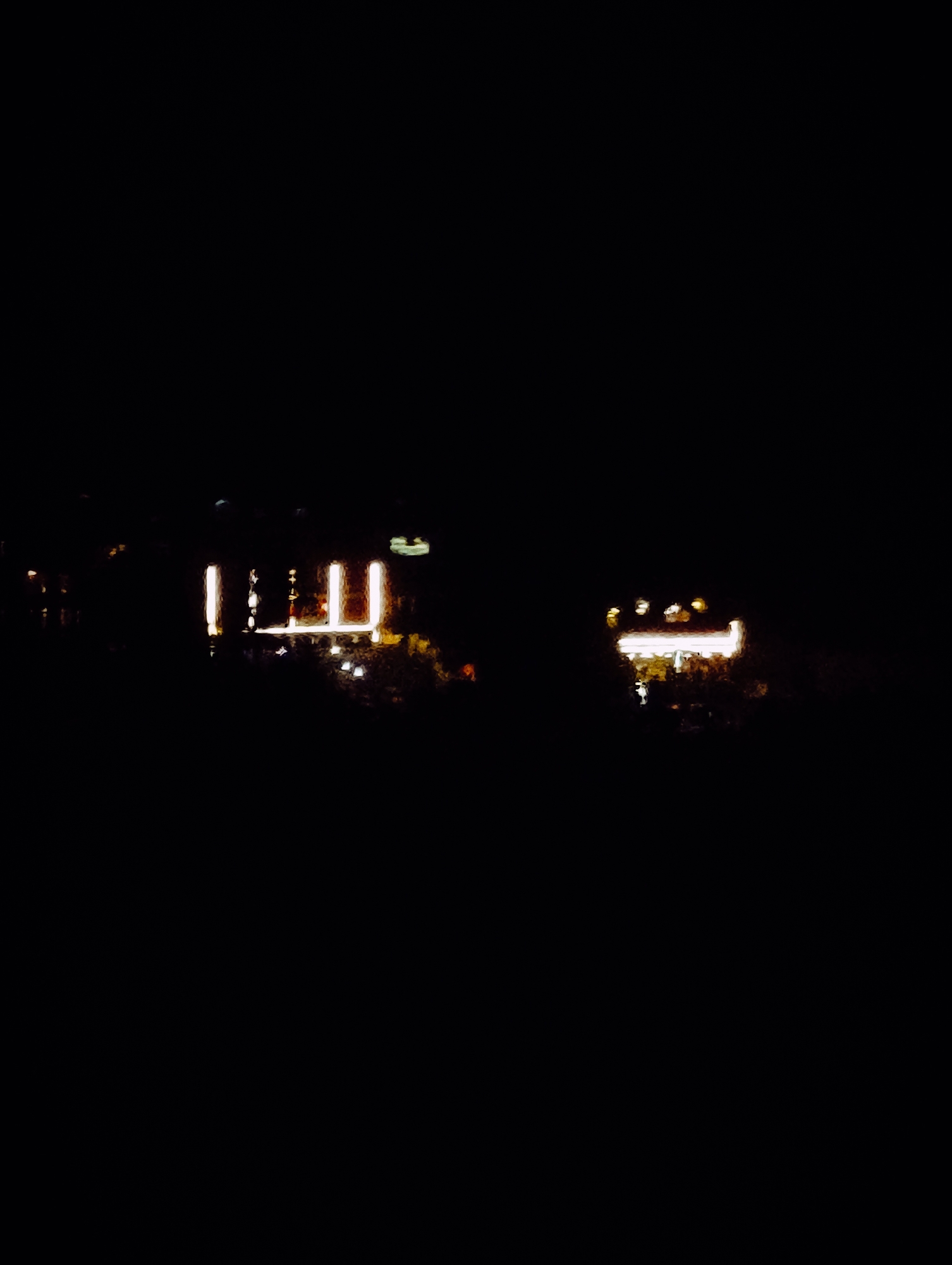 The image is a nighttime photograph taken in Vashisht, India. It shows a dark environment with artificial lights creating blurred shapes and reflections. The primary illuminated objects appear to be buildings or structures with bright windows. In the foreground, there is a dark foreground, and the background is also dark, suggesting a cityscape at night. The lights are hazy and out of focus, giving the image an abstract quality. There are no visible people or specific identifiable landmarks in the image.