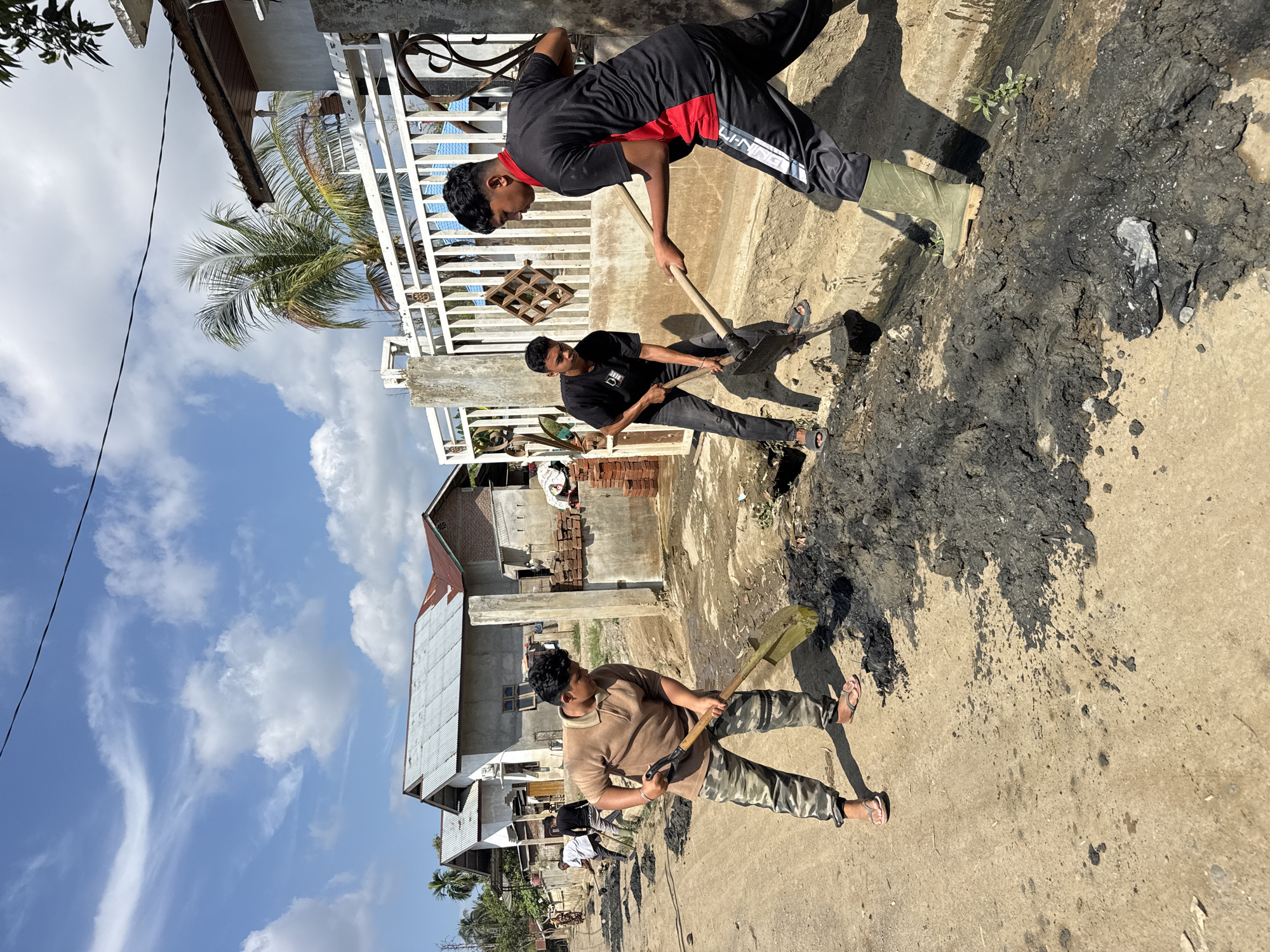 In Meunasah Raya, Indonesia, four young men are engaged in manual labor, clearing debris from a roadside drain. The scene is set on a dusty, unpaved street, with houses and a white metal fence lining the right side of the road. The sky is a bright blue with scattered white clouds.
The primary focus is on the four individuals actively working. The man closest to the viewer, wearing a brown polo shirt and camouflage pants, is using a shovel to dig into a pile of dark, wet mud. He is barefoot, wearing flip-flops. To his left, another man, dressed in a white shirt and dark pants, is also shoveling mud. Further down the street, two other young men are working together. One, in a black t-shirt and dark pants, is using a shovel to move mud, while the other, in a black and red athletic shirt and black pants, is holding a shovel and appears to be directing or assisting. He is wearing green rubber boots.
The debris they are clearing consists of dark, clumpy mud, which has accumulated in the drain and spilled onto the street. Several piles of this mud are visible. In the background, the houses have corrugated metal roofs, some showing signs of rust. A palm tree can be seen behind the fence. The overall impression is one of community effort in maintaining the local infrastructure.