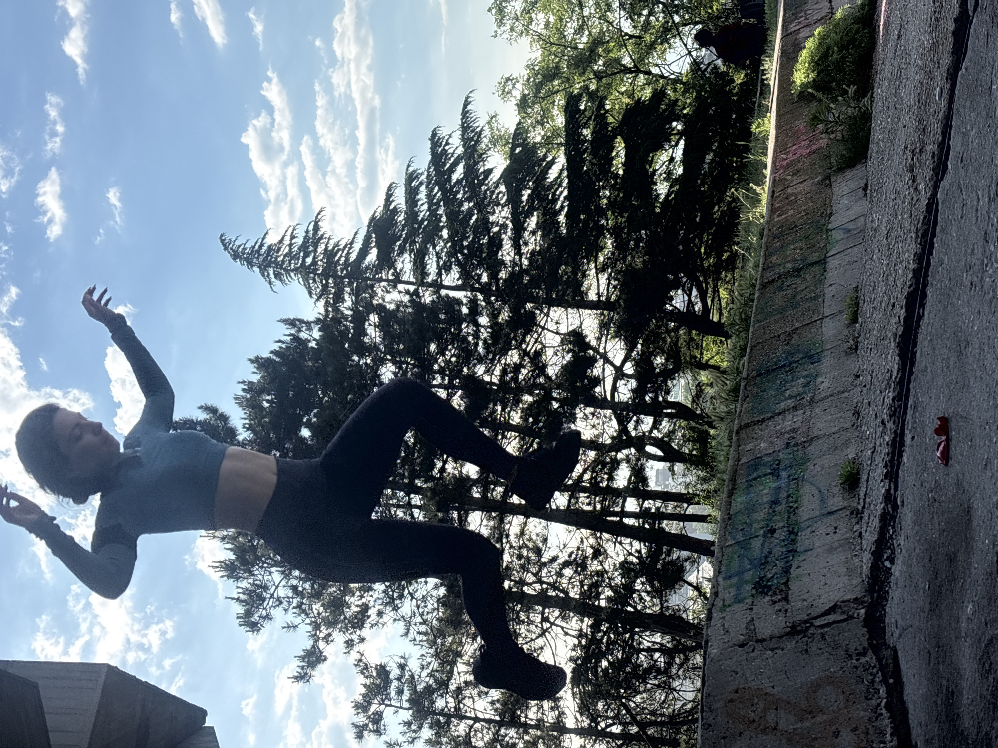 The image is a dynamic photograph, appearing rotated clockwise, capturing a person suspended against a bright sky. A young woman, dressed in a dark long-sleeved athletic crop top, dark leggings, and dark shoes, is in mid-air. Her arms are raised and bent, and her legs are bent at the knees, conveying a powerful and weightless pose, suggesting a jump, fall, or acrobatic movement. Her body is largely silhouetted against the vibrant blue sky, which features scattered white clouds.
The background elements are also rotated, with a dense line of tall, dark evergreen trees silhouetted against the sky and strong sunlight filtering through their branches. To the right of the frame, a textured concrete structure, appearing as a vertical wall or embankment, extends upwards. This weathered concrete has patches of small green vegetation growing on its surface and along its top edge, as well as faint, indecipherable graffiti in blue and green hues. Below this 'wall' and adjacent to it, is a dark, paved surface, where a pair of red sunglasses is visible.
The bright, clear daylight suggests it is mid-morning or afternoon. The combination of natural elements like trees and sky with urban or semi-urban infrastructure like the concrete wall and pavement indicates an outdoor setting, consistent with Varna, Bulgaria. The image evokes a strong sense of dynamic movement and freedom.