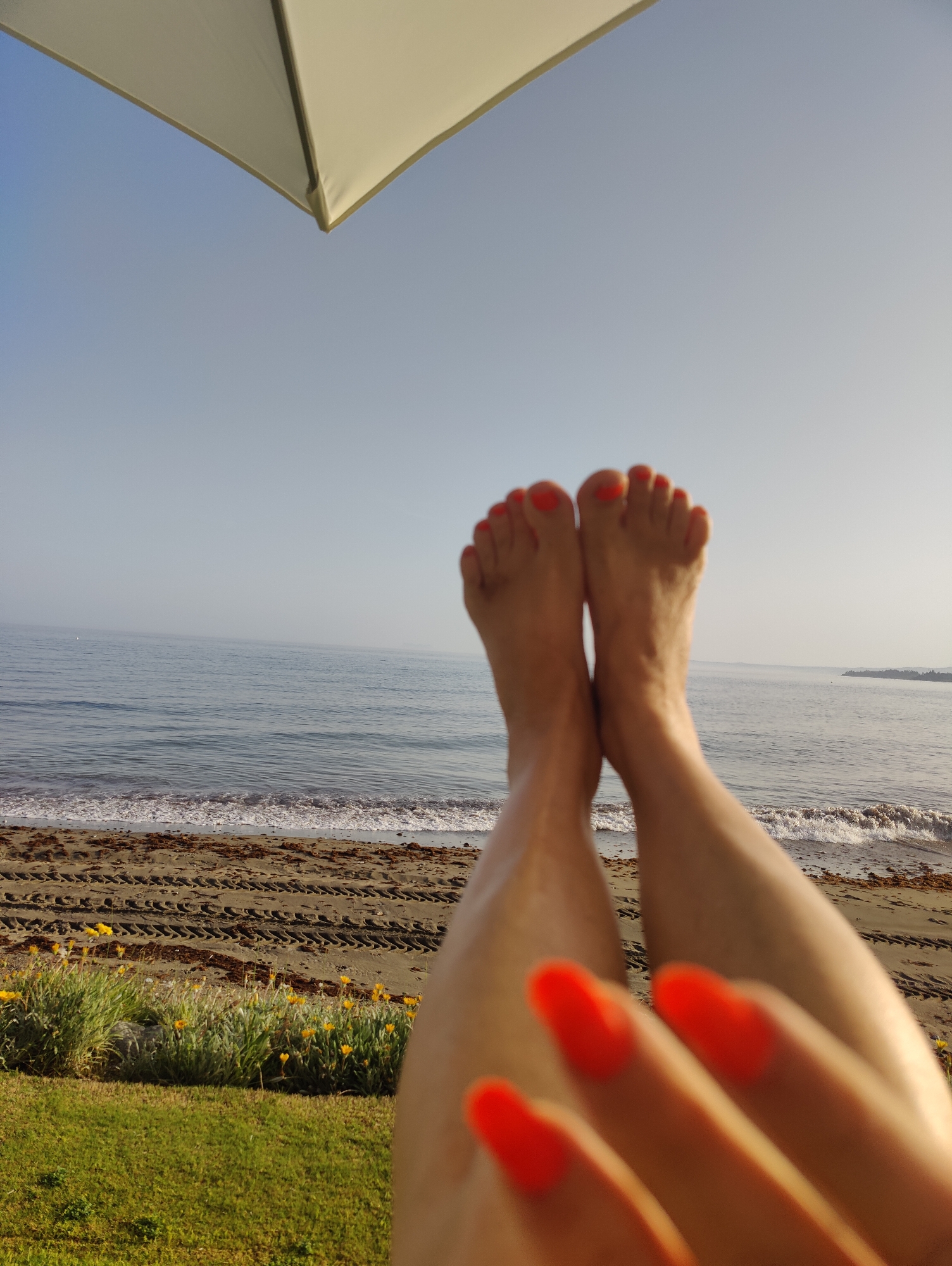 A first-person perspective shot shows a person's bare feet with bright orange nail polish against a beach and ocean backdrop. An umbrella's edge is visible in the upper left corner, casting a shadow. The beach has tire tracks imprinted on the sand, with a line of green grass and yellow flowers in the foreground. The ocean waves are gentle, and the sky is clear with a hint of haze.