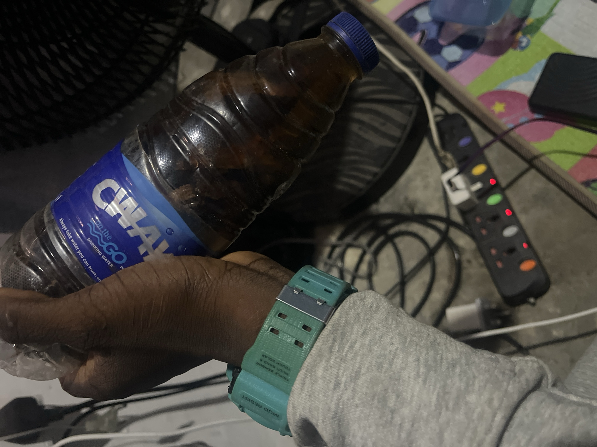 A hand holds a partially consumed bottle of CWAY "on the GO" drinking water. The bottle has a blue cap and the contents appear to be dark liquid, possibly tea or flavored water. The hand is wearing a turquoise-colored watch with a darker grey accent. The arm is covered by a grey sweatshirt. In the background, there is a fan, various cables and plugs, an extension cord, and a colorful patterned mat. The location appears to be indoors. There is no information about the city of Ado, Nigeria.