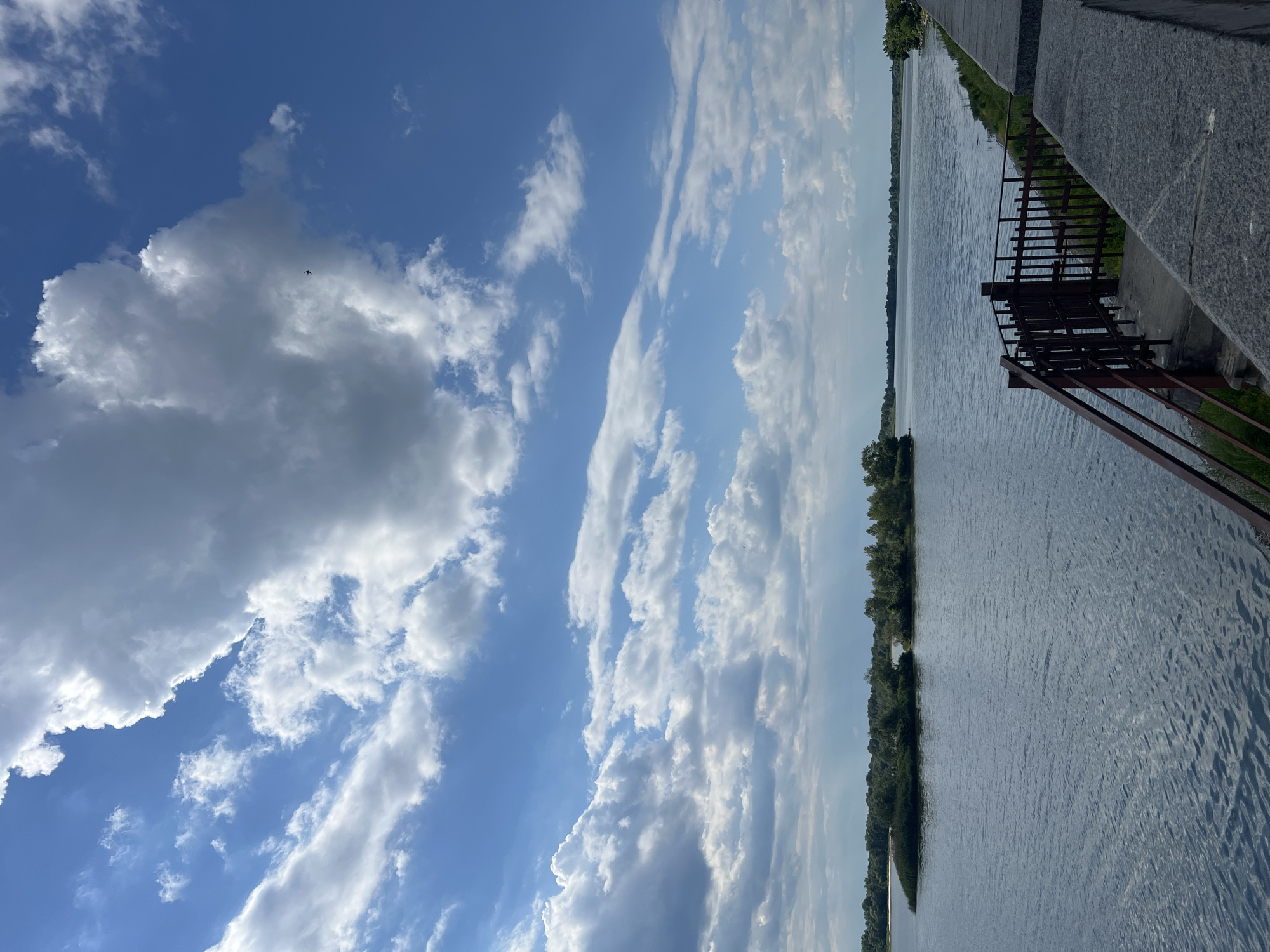 A vertical, eye-level, outdoor shot captures a tranquil scene by a body of water under a bright, cloud-filled sky. The water, a deep, calm blue, reflects the sky and is subtly rippled. In the middle distance, a small, lush green island dotted with trees breaks the expanse of water. To the right, a gray, stone embankment leads the eye toward the foreground, where a weathered metal railing and stairs descend towards the water's edge. The sky above is a vibrant blue, adorned with dramatic white and gray clouds, some billowing and voluminous, others wispy and thin. A tiny black bird is visible in flight against the sky. The scene appears to be a park or waterfront area, likely within a city given the presence of the embankment and railing, though the specific location is unknown. The overall atmosphere is peaceful and serene.