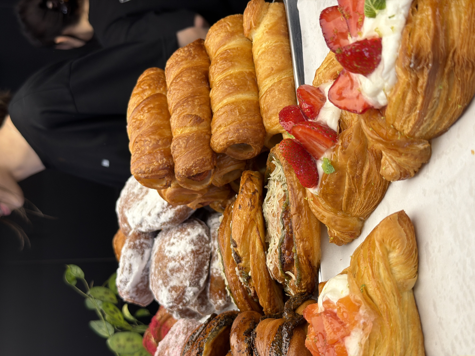 The image shows a close-up of a display of baked goods, likely in a bakery or cafe. There are various pastries, including what appear to be croissants, danishes topped with strawberries and cream, and possibly some savory items. In the background, two individuals wearing black tops are partially visible. The person on the left has their head turned, with their mouth open as if speaking or reacting to something. The person on the right has their back to the camera. The overall lighting suggests an indoor setting. The location of Izhevsk, Russia is not determinable from the image.