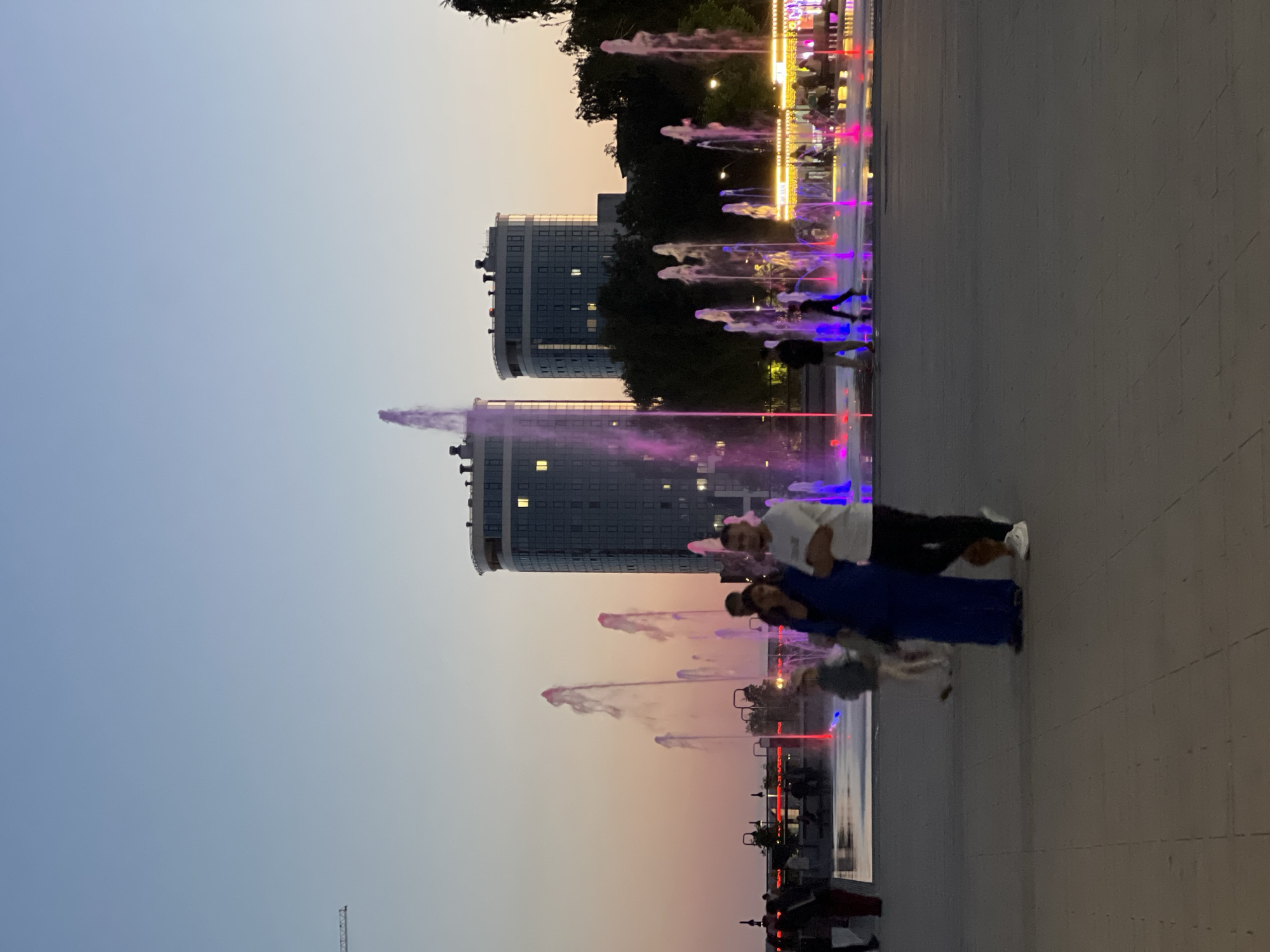 The image shows a view of the Izhevsk National Congress Palace at dusk. A group of people are gathered around a fountain, which is illuminated with colorful lights. The fountain sprays water in arcs, creating a dynamic display. In the background, two modern buildings with many windows stand tall. The sky is a soft gradient of orange and blue, indicating the end of the day. The scene is set in a public square, with paved ground and some trees visible.