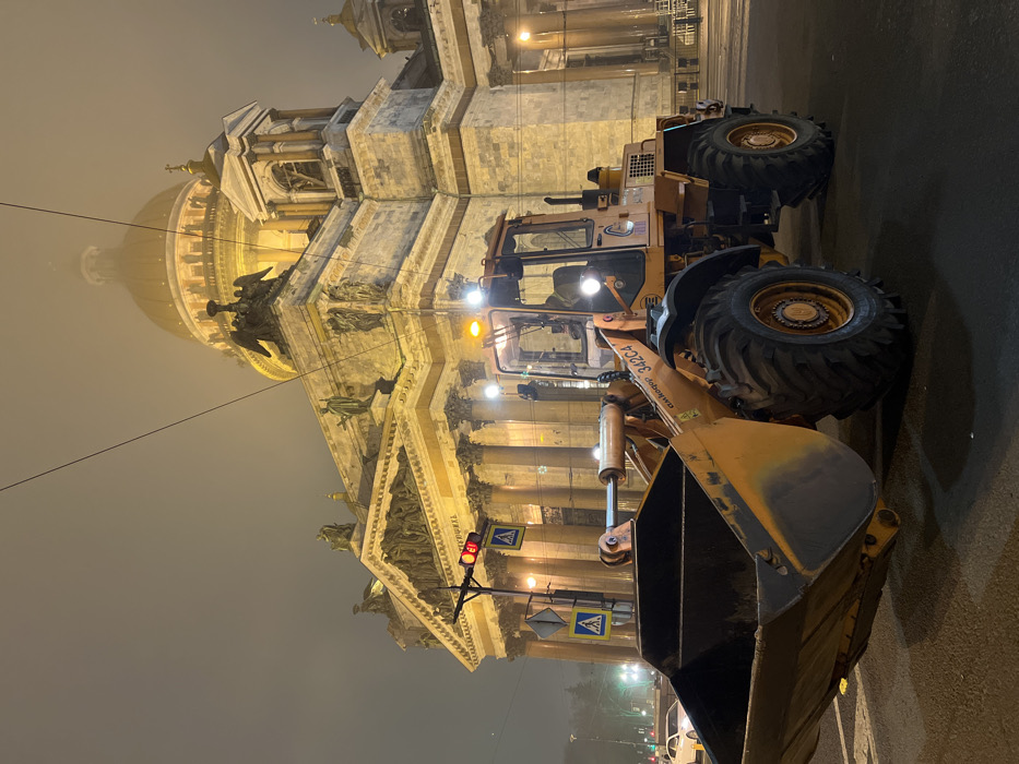 The image depicts an urban night scene in Saint Petersburg, Russia, with Saint Isaac's Cathedral prominently illuminated in the background. In the foreground, an orange Amkodor 342C4 wheel loader is stationary on a dark asphalt road. The loader's headlights and amber warning lights on its roof are illuminated. An individual is visible inside the operator's cab. To the left of the loader, a traffic signal displays a red light, and two blue pedestrian crossing signs are mounted on a pole. Overhead electrical wires traverse the upper portion of the frame. The neoclassical facade of Saint Isaac's Cathedral features granite columns, pediments with sculptures, and rooftop statues, all brightly lit. Its large golden dome is partially obscured by fog or mist, which creates a soft halo effect around the building's upper structure. Distant streetlights and a few indistinct vehicles are visible further down the street to the left. The overall atmosphere is dark and foggy, illuminated by artificial light sources.