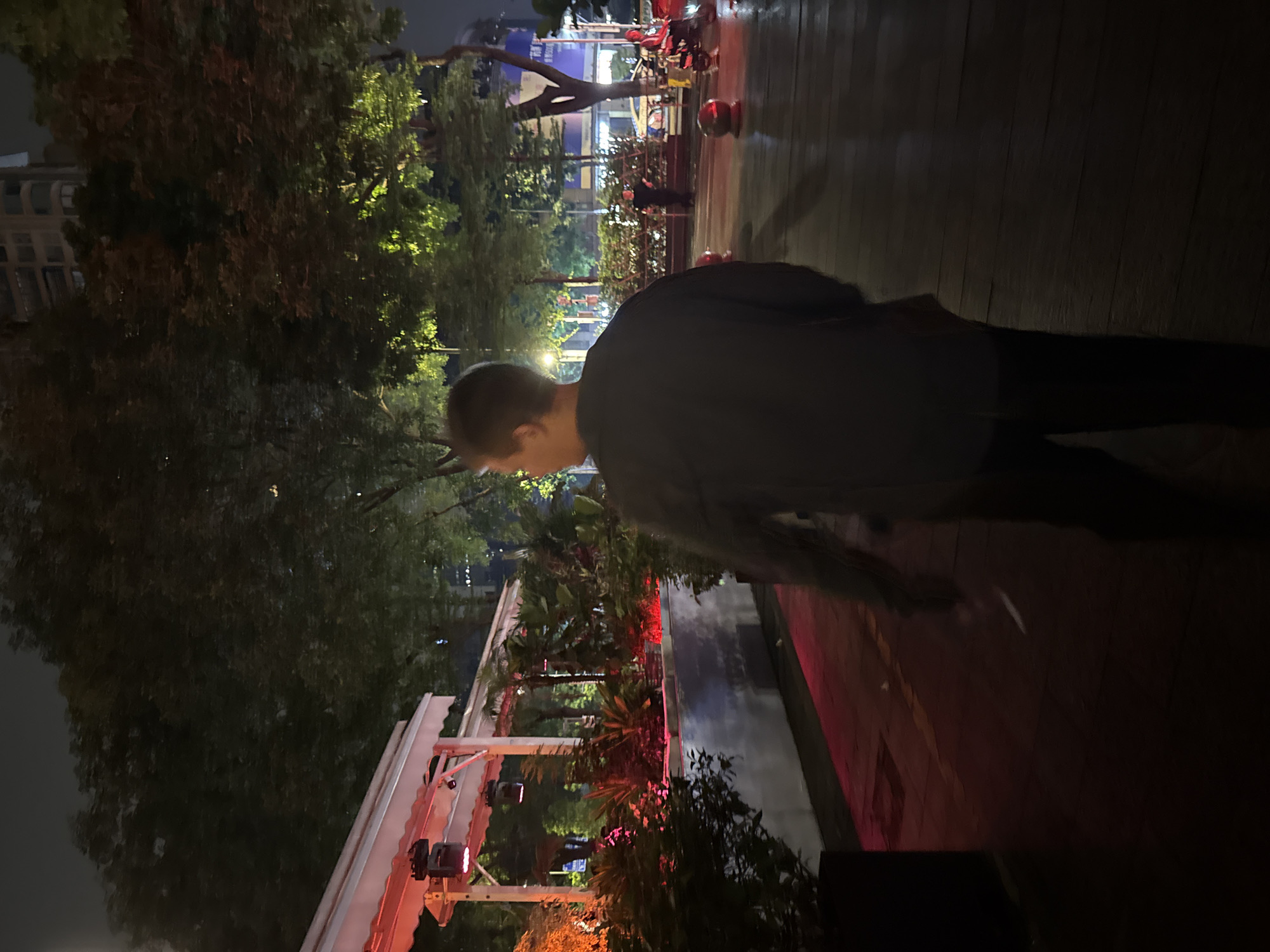 The image shows an outdoor scene at night. A person with short hair is walking on a walkway, with trees and buildings in the background. There are red lights illuminating plants and structures. Based on the location cues and visual characteristics, the scene is plausibly located in the Tianhe District of China, although the specific location cannot be precisely identified.
