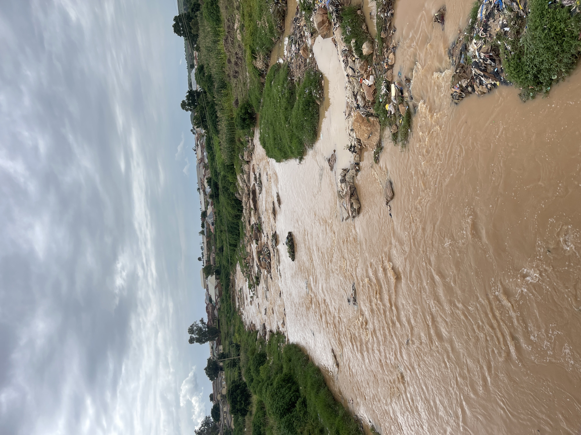 The image depicts a muddy river flowing through a populated area, likely in Rafin Par, Nigeria, under a cloudy sky. The river carries a significant amount of debris, including plastic waste, household garbage, and other refuse, which is accumulated along its banks and in the water. The riverbanks are lined with lush green vegetation, and in the background, there are numerous houses with reddish-brown roofs, indicative of a residential area. The scene suggests an environmental concern related to waste management and pollution impacting the river system.