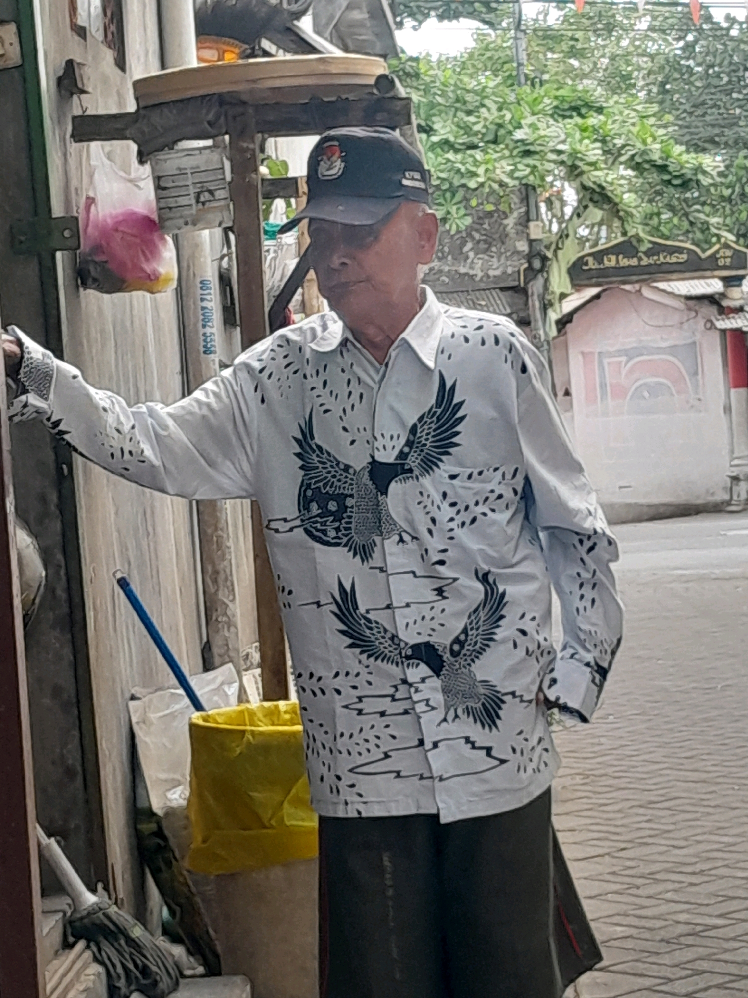 An elderly man wearing a white shirt with a black bird print, a black baseball cap, and dark pants stands near a building. He is leaning against a wall. In the background, there is a street with a brick-paved sidewalk. Trees and a building with Arabic writing are visible. Several objects are near the man, including a yellow trash bag, a mop, and a pink plastic bag filled with items. The overall scene appears to be an outdoor urban environment.