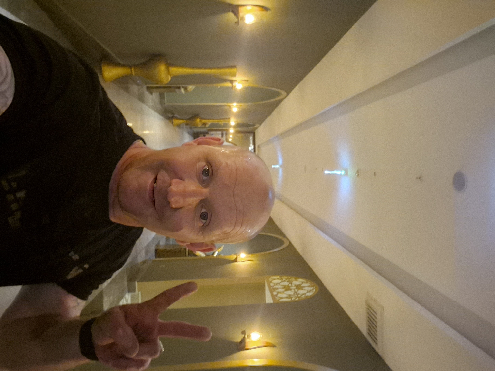 A balding, middle-aged man, wearing a black t-shirt, is making a peace sign with his left hand, with a fitness tracker on his wrist. He is inside a corridor with recessed lighting fixtures along the side walls and on the ceiling. Some decorative, oval cutouts are along one wall. The location is the city of Alanya, Turkey.