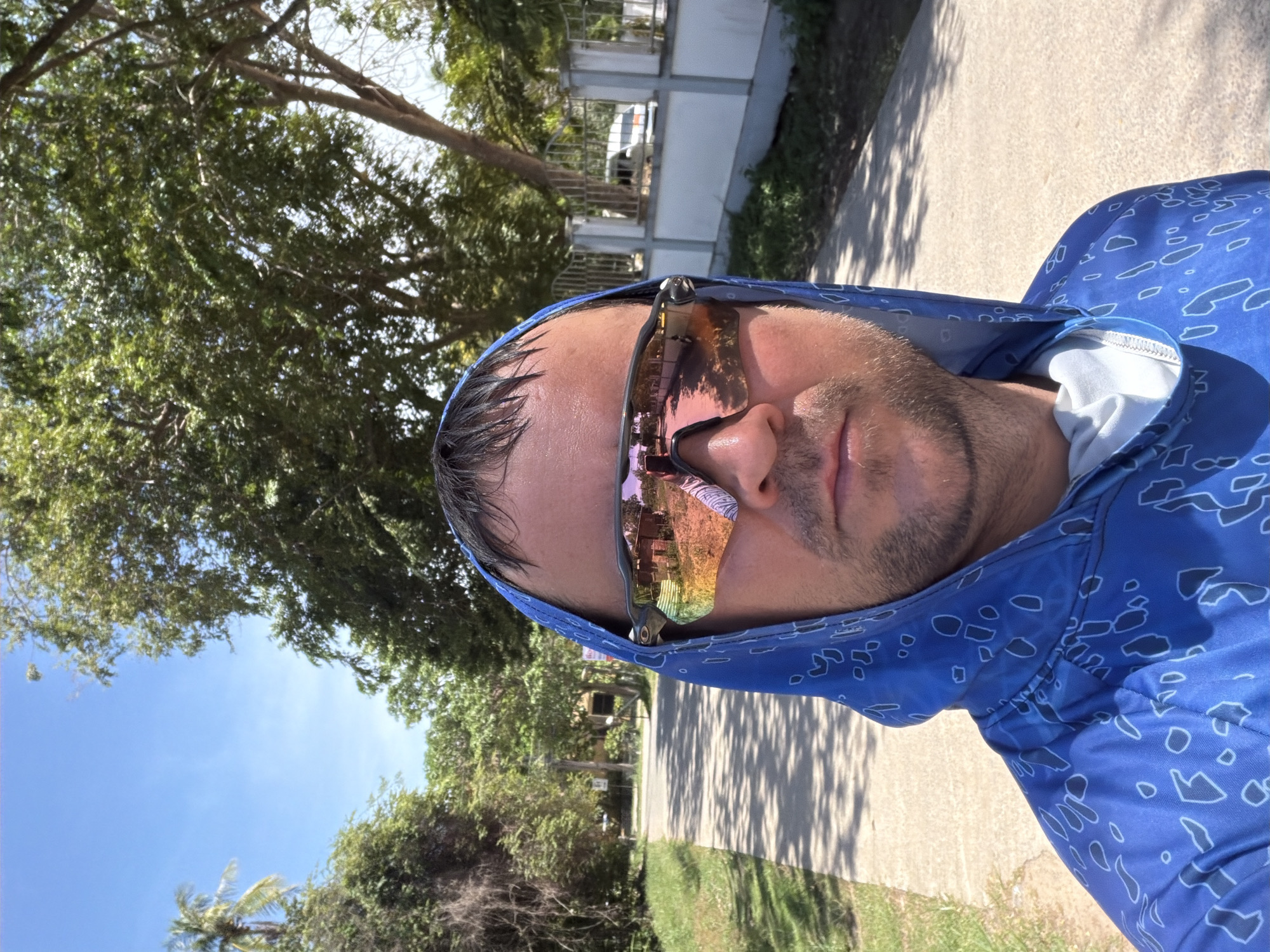 A man wearing a blue hooded shirt with a white pattern is taking a selfie outdoors on a sunny day. He is wearing sunglasses that reflect a scene with buildings and people. The man is standing on a paved walkway lined with grass and trees. In the background, there is a white fence with a white van behind it. The location appears to be Baan Bo Phut, Thailand, given the tropical vegetation and architectural style.