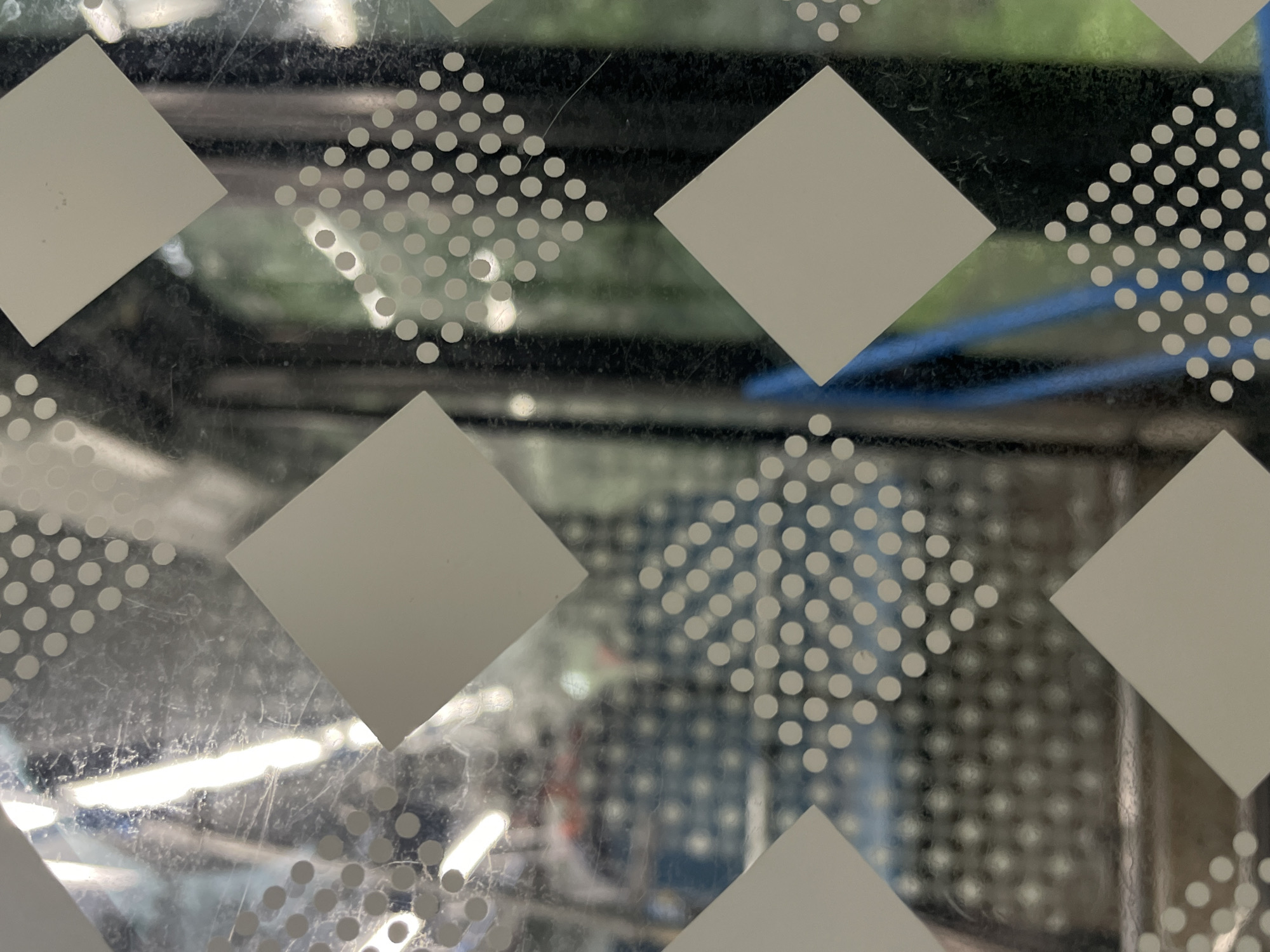 The image displays a close-up view of a glass surface with a diamond-patterned design. The pattern consists of large, solid white diamonds interspersed with clusters of small white dots arranged in diamond shapes. The glass appears to be part of a window or screen, as a blurred background is visible through it. The background shows hints of an indoor or outdoor environment with various objects, including what seem to be shelves, possibly containing items, and some greenery, suggesting a setting like a store, greenhouse, or an area with plants. There are also horizontal lines and a blue element in the background, which could be part of shelving or furniture. The lighting suggests it is daytime, with some bright reflections on the glass, indicating artificial or natural light sources. The overall scene is abstract due to the focus on the patterned glass and the blurred background. There is no discernible text in the image.