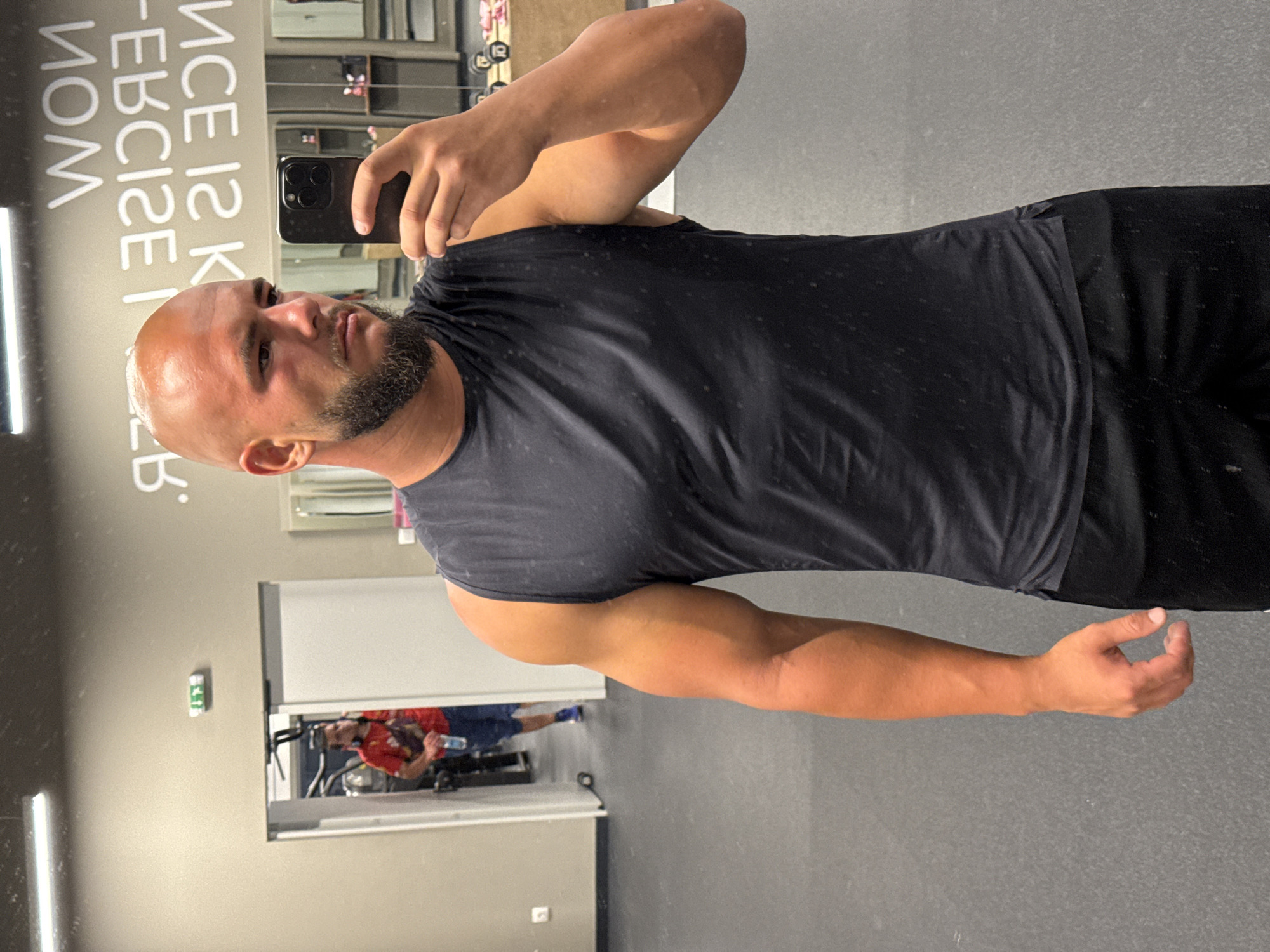 A man is taking a mirror selfie in a gym. He is bald with a beard and is wearing a dark grey sleeveless t-shirt and black shorts. He is holding a black smartphone in his right hand. In the background, another man wearing a red t-shirt and blue shorts is visible through an open doorway, holding a water bottle and wearing headphones. The gym has a grey floor and walls with motivational text visible on the wall behind the selfie-taker, reading "WON EXERCISE IS KICKING". There are weight racks and mirrors in the background. The scene is indoors. The time of day is not discernible. The location is implied to be in Sofia, Bulgaria, based on the provided information.