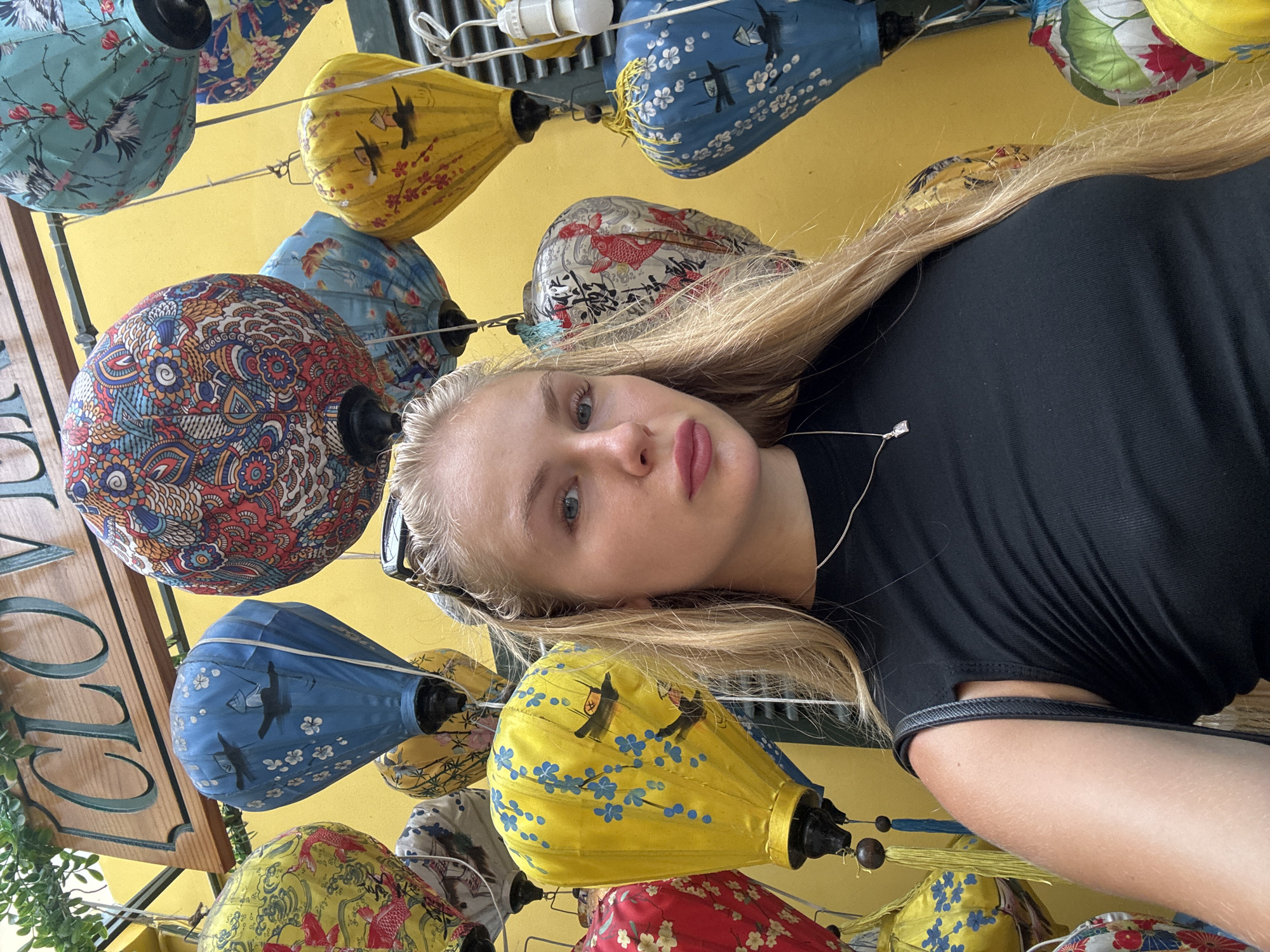 This is a selfie taken by a young woman with blonde hair, wearing a black sleeveless top and a delicate necklace. She is standing in front of a wall covered with colorful Vietnamese lanterns, many of which are patterned with traditional designs. A wooden sign with the word "CLOVER" is visible in the upper left corner. The vibrant lanterns and the yellow wall suggest a location in a bustling marketplace or tourist area, possibly a shop selling traditional Vietnamese crafts. The overall scene is bright and cheerful, indicative of a daytime setting. While the exact location in Nha Trang is not specified, the presence of traditional lanterns and the vibrant atmosphere are characteristic of tourist-friendly areas in Vietnamese cities.
