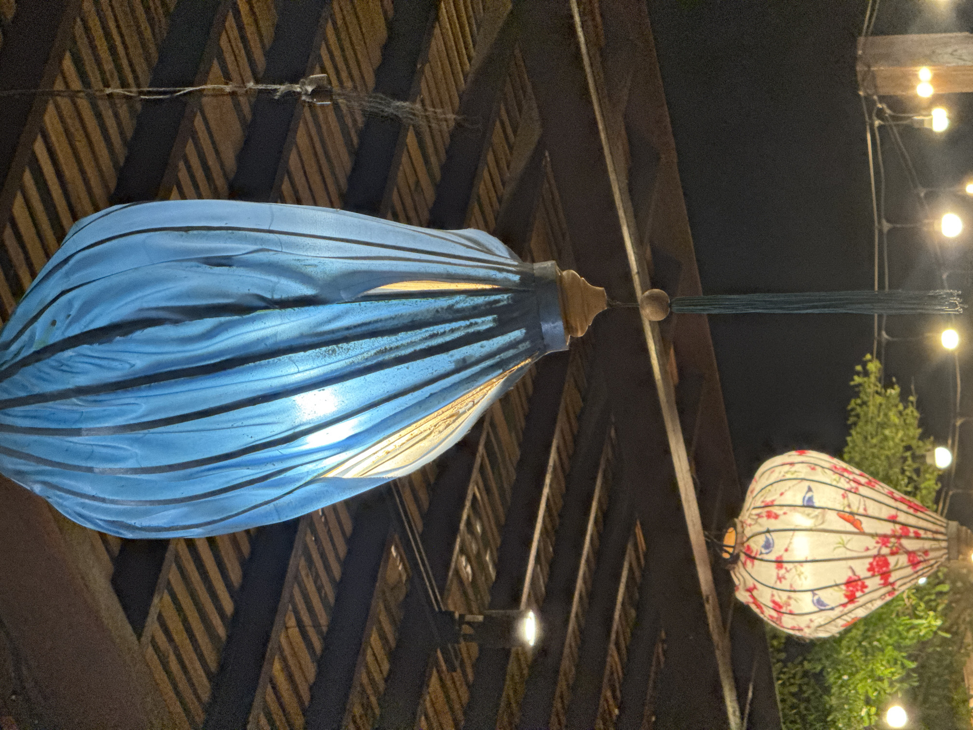 The image captures an atmospheric nighttime scene in Nusa Penida, Indonesia, featuring decorative lighting and a rustic wooden structure. In the foreground, a prominent, elongated blue lantern hangs, made of translucent, pleated fabric with distinct dark horizontal lines. It glows brightly from an internal light source, illuminating its textured surface, and is adorned with a wooden finial and a tassel of blue threads at its base.
Above and partially behind the blue lantern, a strong wooden beam or trellis structure is visible, forming a prominent diagonal pattern and casting sharp shadows. A thin string or wire runs across these wooden elements.
Further into the background, to the right, another traditional-style lantern is visible. This lantern is off-white with intricate red floral patterns and small blue bird motifs, complementing the festive ambiance. Behind this patterned lantern, green foliage from a tree is discernible against the dark night sky. A string of warm, glowing fairy lights stretches across the upper right portion of the frame, enhancing the cozy and inviting atmosphere.
The scene is set outdoors at night, indicated by the dark sky and the exclusive reliance on artificial illumination from the lanterns and string lights. The traditional design of the lanterns is consistent with the cultural context of Indonesia. No specific activity is taking place, and there is no visible text.