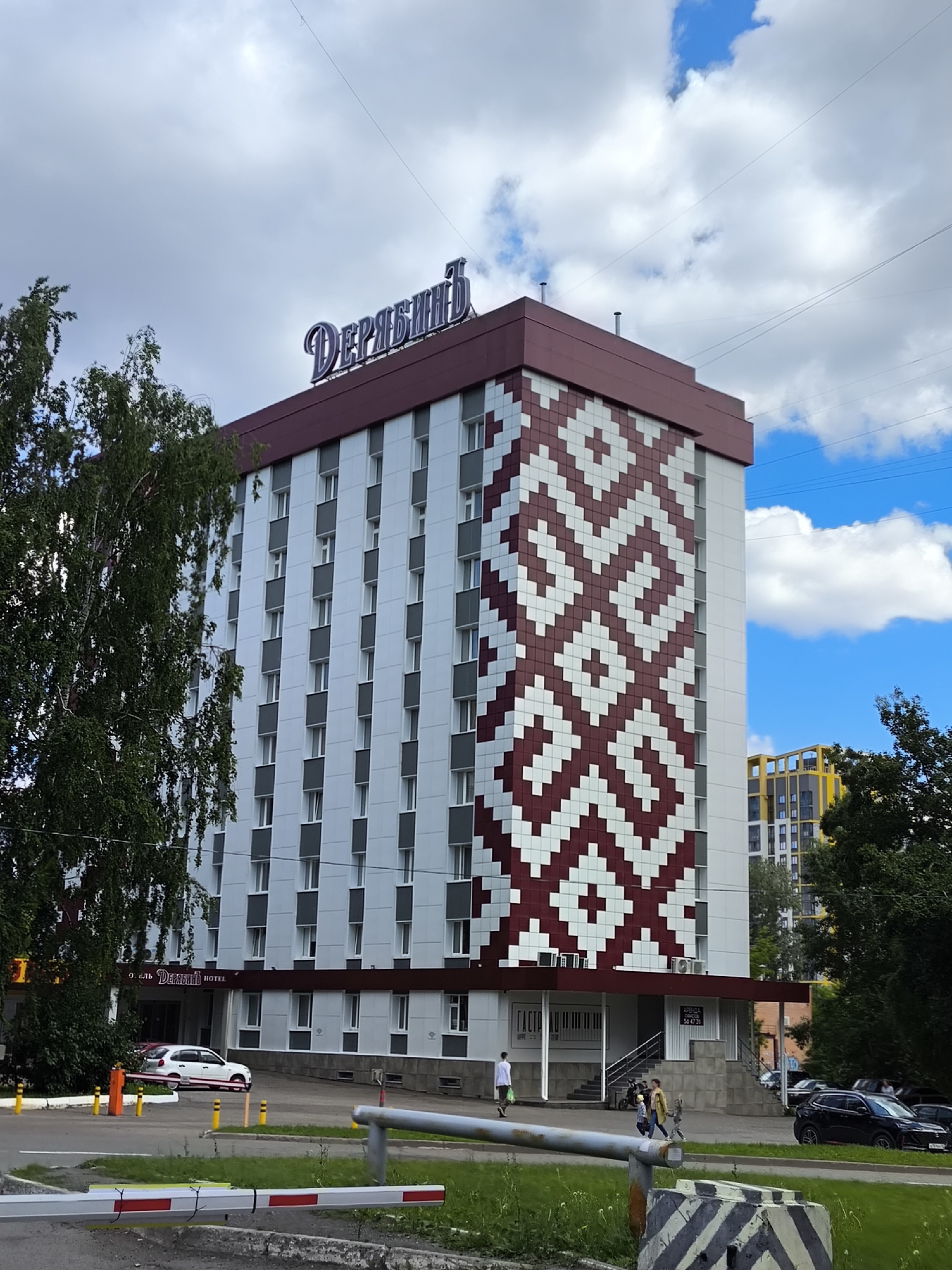 The image depicts the Deryabin Hotel, located in Izhevsk, Russia, under a partly cloudy sky. The hotel is a multi-story building, approximately 8-9 floors high, featuring a distinctive architectural design. The main facade, visible on the right, is adorned with a large, repeating geometric pattern composed of maroon and white squares, resembling a traditional woven design. The other visible facades are predominantly white with grey horizontal bands beneath the windows. The building's roofline and lower sections are finished in a dark red or maroon color.
Prominently displayed on the roof is a large, stylized sign spelling "ДерябинЪ" (Deryabin). Another sign above the main entrance reads "ОТЕЛЬ ДерябинЪ HOTEL." Near the ground level on the right side, a partially visible sign indicates "ГАСТРО..." (Gastro...) and another sign above steps advertises "АРЕНДА" (Rent) with a phone number "56 47 31."
In the foreground, a white and red striped barrier crosses a paved area on the left, with several yellow and orange bollards on a grassy strip beside it. A grey and black striped concrete block is visible in the bottom right corner. A large, leafy tree partially obscures the left side of the hotel.
Several individuals are present on the paved area around the hotel's entrance. One person in a white shirt walks away from the main entrance on the left. Another person in a light-colored jacket walks with a small child near the "АРЕНДА" sign on the right.
Vehicles include a white car with pink wheel accents parked near the hotel entrance on the left and a black SUV parked further down the street on the right. In the background to the right, a tall, modern residential building with yellow and grey sections is visible. The sky features a mix of white clouds and patches of clear blue, with thin utility lines crisscrossing overhead.