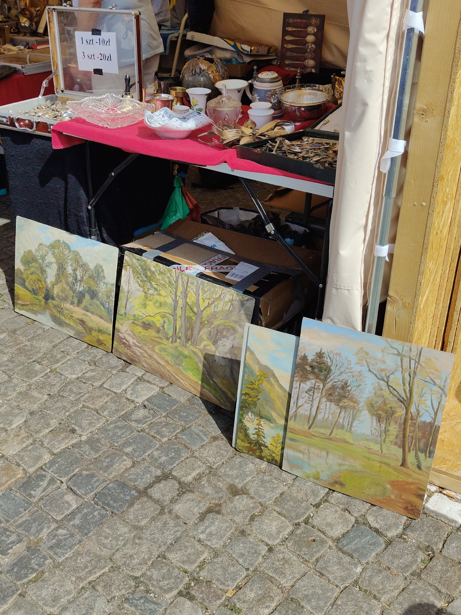 Four landscape paintings are displayed on a cobblestone surface in front of a market stall. The paintings depict trees, water, and dirt paths, with the closest painting on the right signed by "Zdzislaw N." In the background, a table is covered with a red cloth and displays various items, including a glass bowl, a white bowl, and a tray filled with silverware. A sign with Polish text, possibly pricing information, is visible on a glass display. A person in the background appears to be browsing the items. The market stall is partially covered by a beige awning. The scene is outdoors, likely during daytime, given the shadows cast by the paintings and stall.