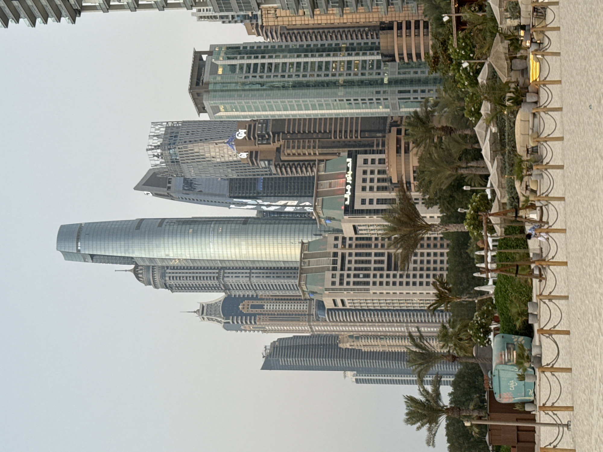 The image depicts a beachfront area with a backdrop of a dense collection of modern skyscrapers in Dubai Marina, United Arab Emirates. In the foreground, a sandy beach is visible with a rope barrier and wooden posts. Further back, there is a seating area with cushioned sofas and umbrellas, surrounded by greenery and palm trees. A turquoise-colored food cart with floral designs and cacti is also present. In the middle ground, several people are seen enjoying the beachside amenities, including a person on a swing. The dominant feature of the background is the impressive array of high-rise buildings, characteristic of Dubai's iconic skyline. The sky is overcast, creating a soft, diffused light.