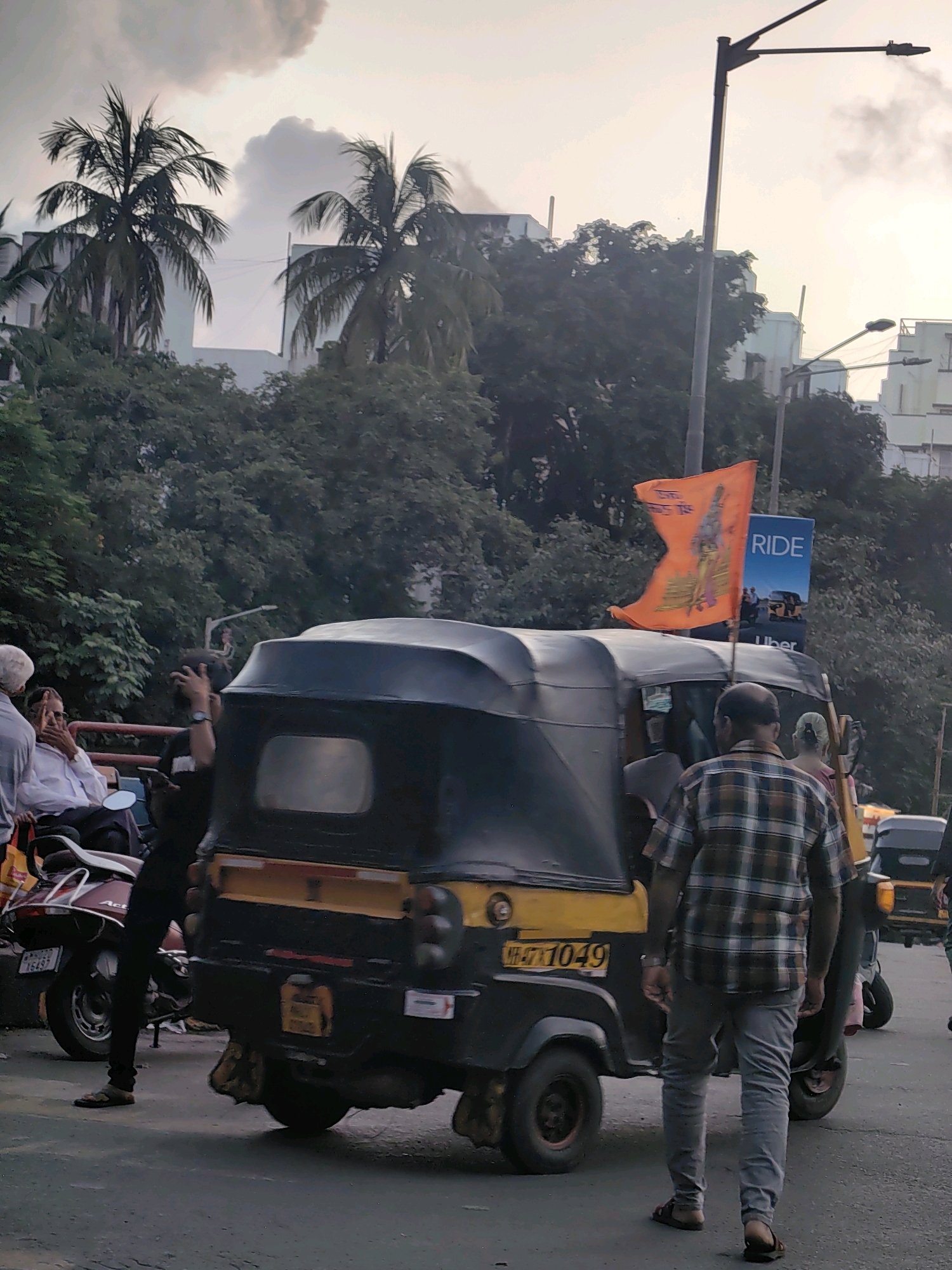 This is a medium shot, eye-level, outdoor, daytime, street-level photograph taken in Mumbai, India. The scene depicts a busy street with several vehicles and pedestrians. In the foreground, a yellow and black auto-rickshaw is parked. The auto-rickshaw has a black canopy and a yellow stripe along its side. It displays a license plate that reads "MH-07-1049". A person wearing a plaid shirt and grey pants is walking away from the camera, towards the right side of the frame. Behind the auto-rickshaw, an orange flag with a depiction of a deity or historical figure is raised on a pole. To the right of the flag, a blue billboard with the word "RIDE" and the Uber logo is visible. In the background, lush green trees obscure buildings. A person is seated on a scooter on the left side of the auto-rickshaw. They are wearing a white shirt and dark trousers. Another person is standing next to the scooter, holding a mobile phone. The sky is overcast with hints of clouds. The overall atmosphere suggests an active urban environment.