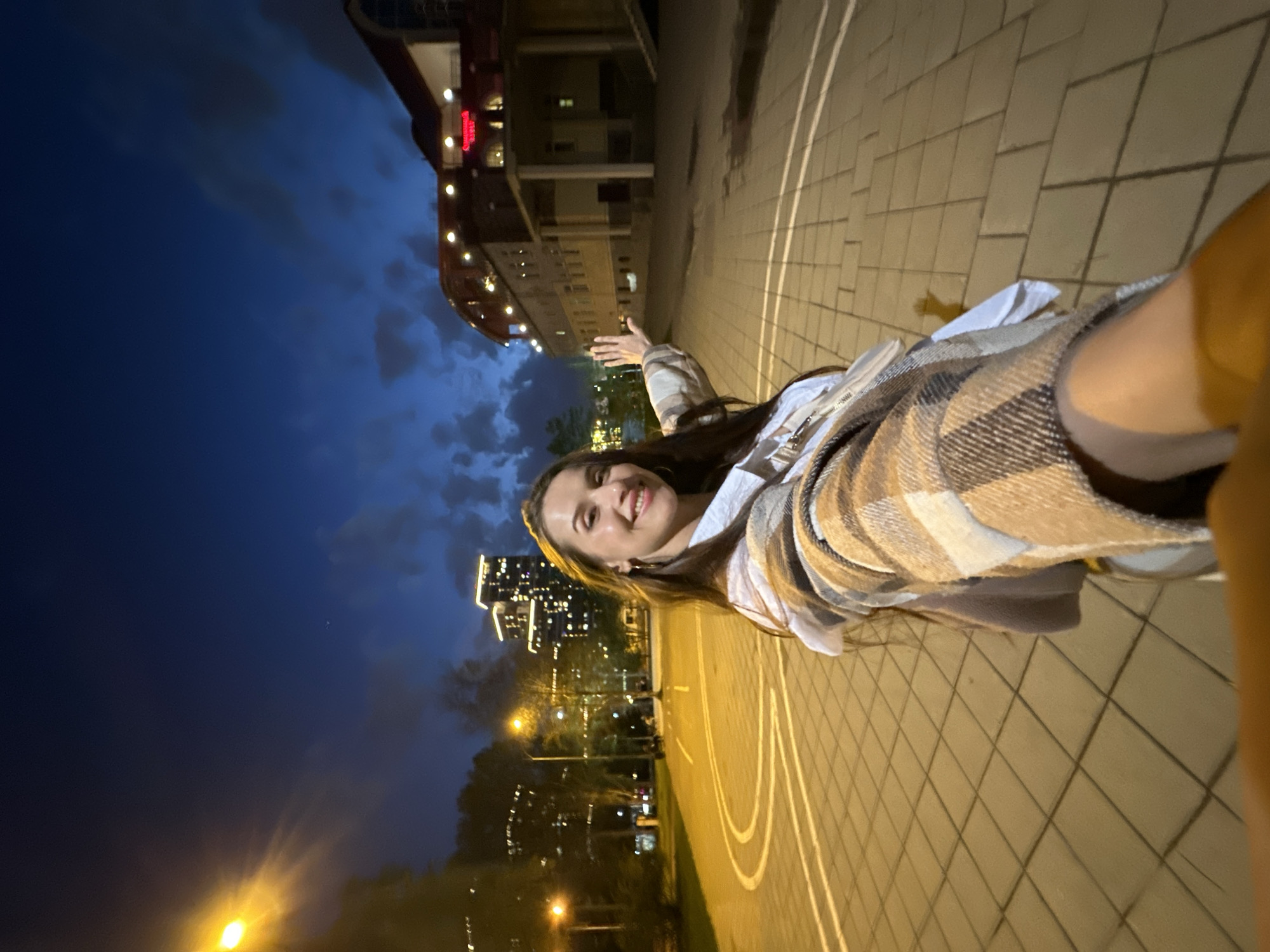 A smiling young woman stands on a basketball court in Yekaterinburg, Russia. It is night, and the city lights are visible in the background, along with a building with illuminated signage. The woman wears a plaid jacket over a light-colored top. She is posing for the camera and has one arm outstretched. The basketball court is tiled with white lines marking the court boundaries.