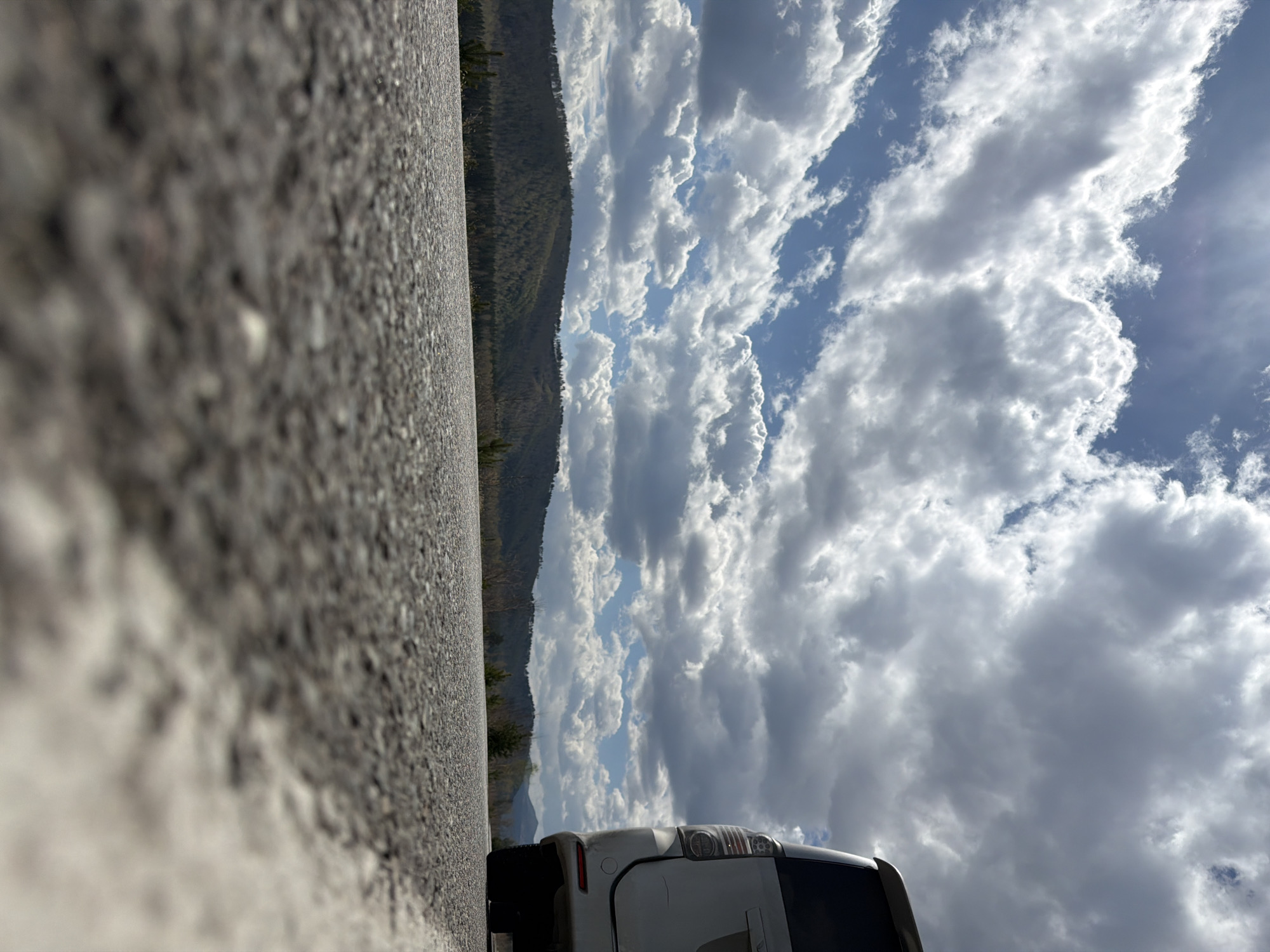 The image depicts a low-angle perspective of a scene that includes a road, a distant mountain range, a cloudy sky, and part of a white car. The foreground is dominated by a coarse, asphalt road surface. A mountain range with dense green vegetation stretches horizontally across the midground. The sky is filled with fluffy white clouds with patches of blue visible in between. The rear of a white car is visible. Based on the current image data, no conclusions can be made about Сизая, Russia.