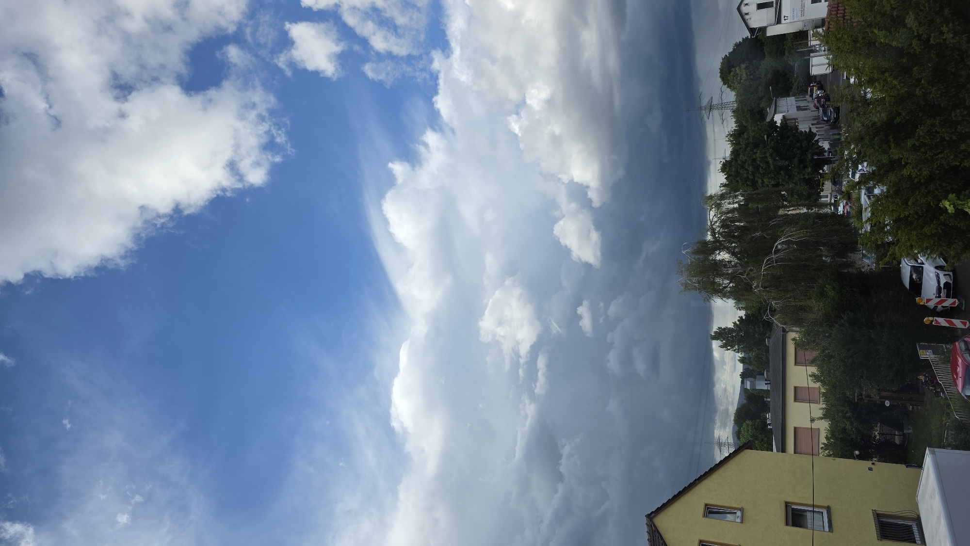 A vertical, slightly downward-angled, eye-level shot captures a residential area under a dynamic sky. The upper half of the image is dominated by a dramatic sky, transitioning from a clear, bright blue with wispy cirrus clouds at the top to a heavily overcast, dark gray expanse of cumulonimbus clouds towards the horizon. The transition zone shows fluffy white cumulus clouds interspersed with darker, more ominous formations, suggesting the potential for rain.
The lower half of the image depicts a street scene with residential buildings and dense greenery. On the left, a two-story, yellow house with a brown roof is prominently featured, showing windows and a small balcony. Adjacent to it are other residential buildings with tiled roofs, some with shutters. A large, mature tree with drooping branches stands in the middle ground, partially obscuring the buildings behind it.
A street is visible running horizontally across the lower third of the frame, lined with parked cars. A white car is parked on the right side of the street, and other vehicles, including a red car, are partially visible. Construction barriers with orange and white stripes are placed near the parked cars, indicating potential road work. Power lines and pylons are visible in the distance, rising above the trees and buildings against the stormy sky. The overall impression is one of a residential neighborhood on a day with changing weather conditions, ranging from clear skies to impending storm clouds.