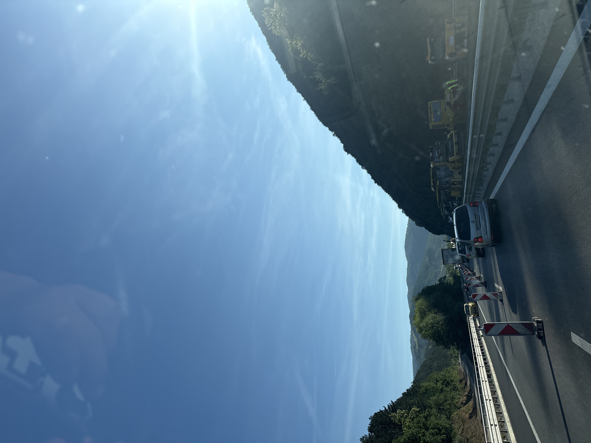 The image captures a highway scene under a clear blue sky with a bright sun on the right. The perspective is from inside a vehicle, looking forward. The highway appears to be under construction or repair, indicated by the presence of traffic cones and various construction vehicles, including yellow rollers and excavators, parked on the right shoulder.
Traffic is visible on the highway, with cars and a truck in the distance. The road has multiple lanes, separated by white lines and a metal guardrail. On the left side of the highway, a dense forest covers a hillside. The right side of the highway is bordered by a steep, vegetated mountain slope. The overall environment suggests a rural or mountainous area.
The image also shows reflections on the vehicle's window, including what appears to be a hand and part of the interior.
While the description includes elements of a highway, construction, and natural surroundings, there is no specific information within the image itself to pinpoint the location to the city of Churek, Bulgaria. The prompt requests a description that includes this location. Without further information or visual cues linking the image to Churek, Bulgaria, this detail cannot be substantiated from the image alone.