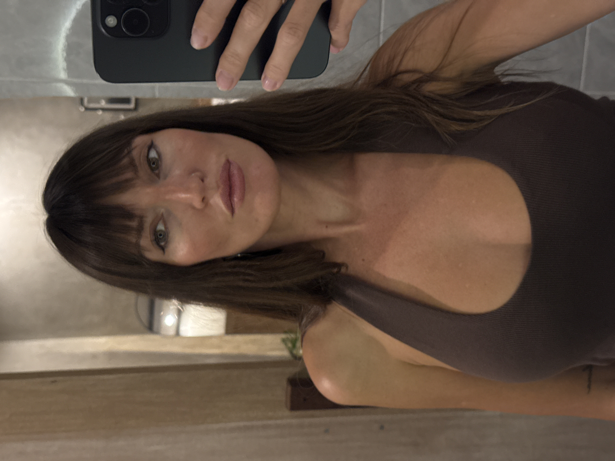 A woman with brown hair and bangs, wearing a brown tank top, is taking a selfie indoors. She is holding a smartphone with a black case. The background is a neutral-toned wall. The location is North Kuta, Indonesia.