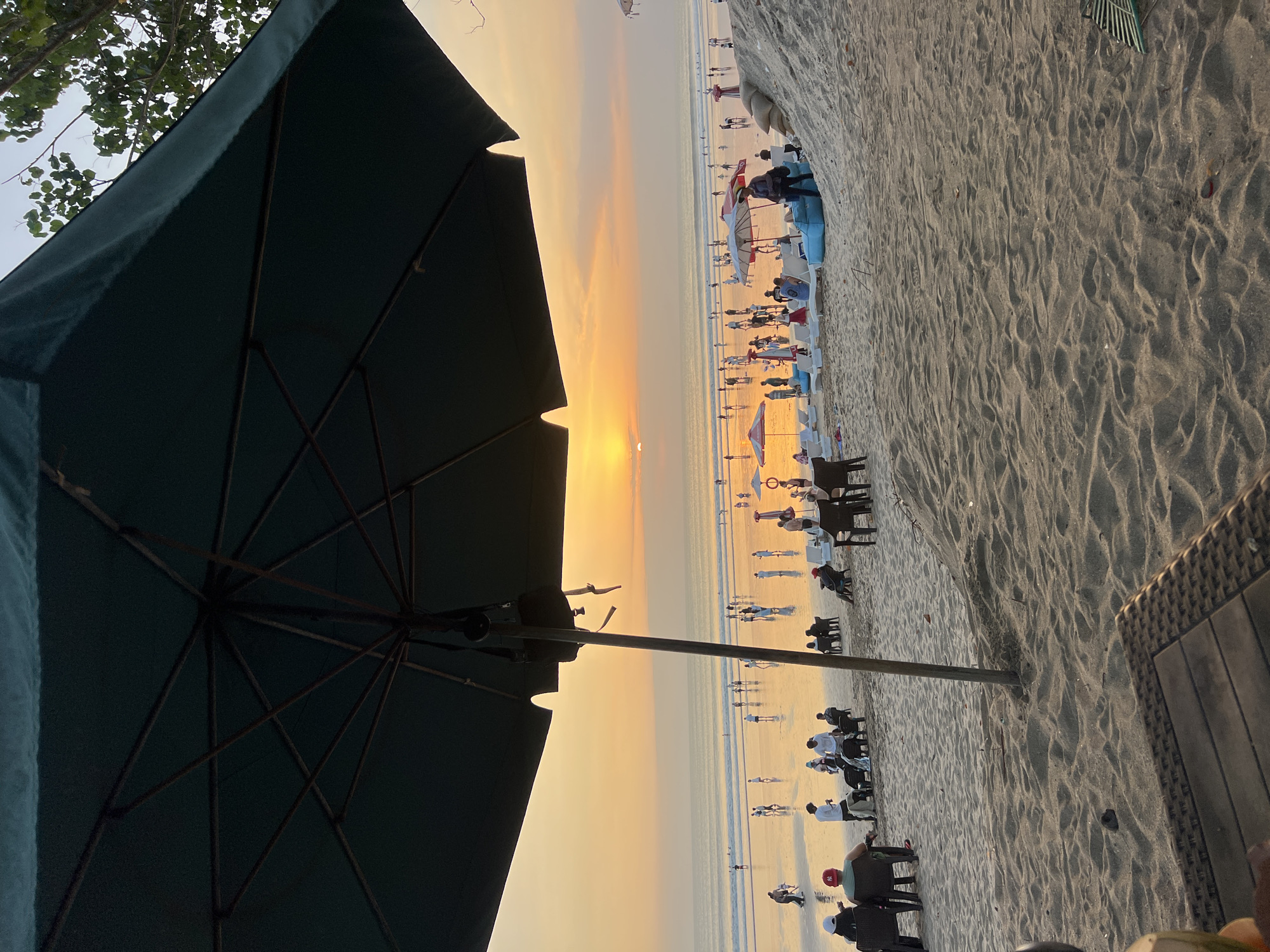 A wide shot captures a serene sunset scene on Kuta Beach, Indonesia. The foreground is dominated by the dark silhouette of a large beach umbrella, its metal ribs subtly visible against the warm, orange glow of the sky. The sand in the immediate foreground is textured, with the gentle undulations of footprints and the rough weave of a woven table visible at the bottom edge of the frame.
Beyond the umbrella, the beach stretches out, dotted with numerous people engaged in various activities. Some are seated in chairs, enjoying the view. Others are strolling along the water's edge, their figures casting long shadows on the wet sand. The ocean itself is calm, reflecting the vibrant hues of the sunset. Several beach umbrellas and lounge chairs are scattered across the sand, indicating a popular gathering spot. The overall atmosphere is tranquil, with the setting sun casting a peaceful ambiance over the beach.