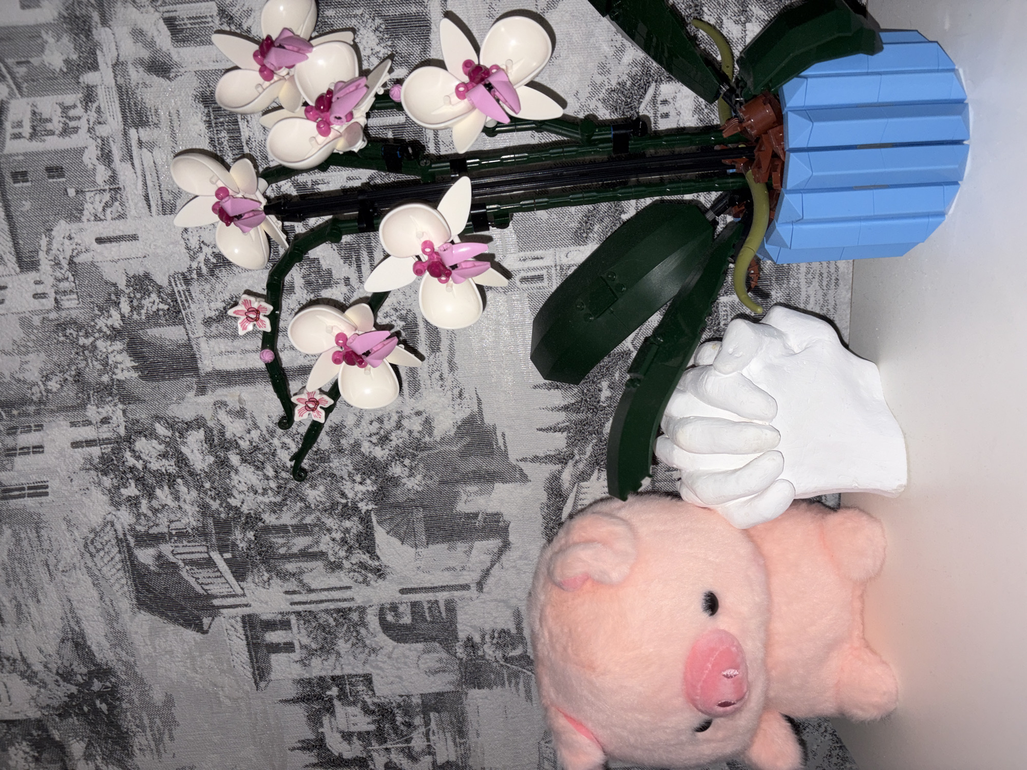 The image shows a still life arrangement. An artificial orchid, likely made of plastic or LEGO bricks, with white petals and pink accents is featured in a light blue pot. Adjacent to the plant is a white plaster hand sculpture. A pink stuffed pig toy rests next to the hand. The arrangement is positioned on a surface covered with a gray patterned cloth, possibly a tablecloth or blanket. The background features a repeating pattern resembling a cityscape or a village, but it is difficult to definitively identify as being in Новинки, Russia. The overall lighting is somewhat bright, with highlights on the surfaces. The scene is static.