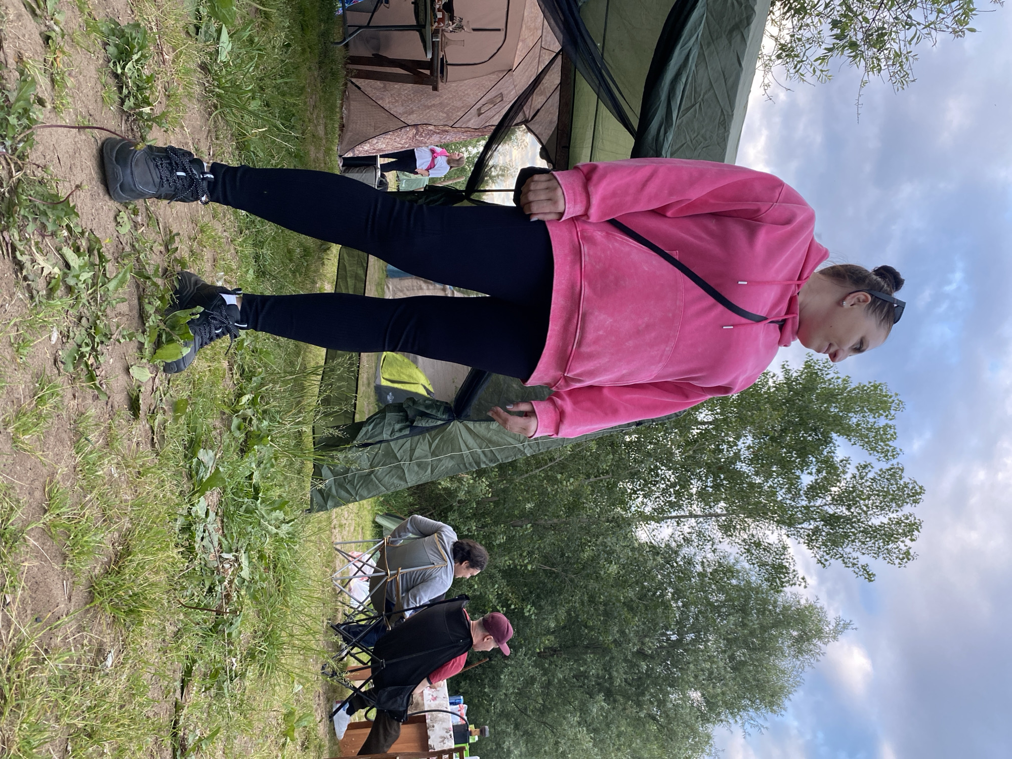 An outdoor scene depicts a woman in the foreground, standing and looking to her right. She is wearing a pink hooded sweatshirt, black leggings, and black sneakers with white laces. A black strap from a bag is visible across her chest. Behind her, a large green tent is partially set up, and a smaller tan tent is visible to the left, with a table in front of it. Further in the background, two men are seated in camping chairs near a table. The environment is a grassy area with trees and a cloudy sky. The image appears to be taken at a campsite. The location is described as Noviy, Russia.