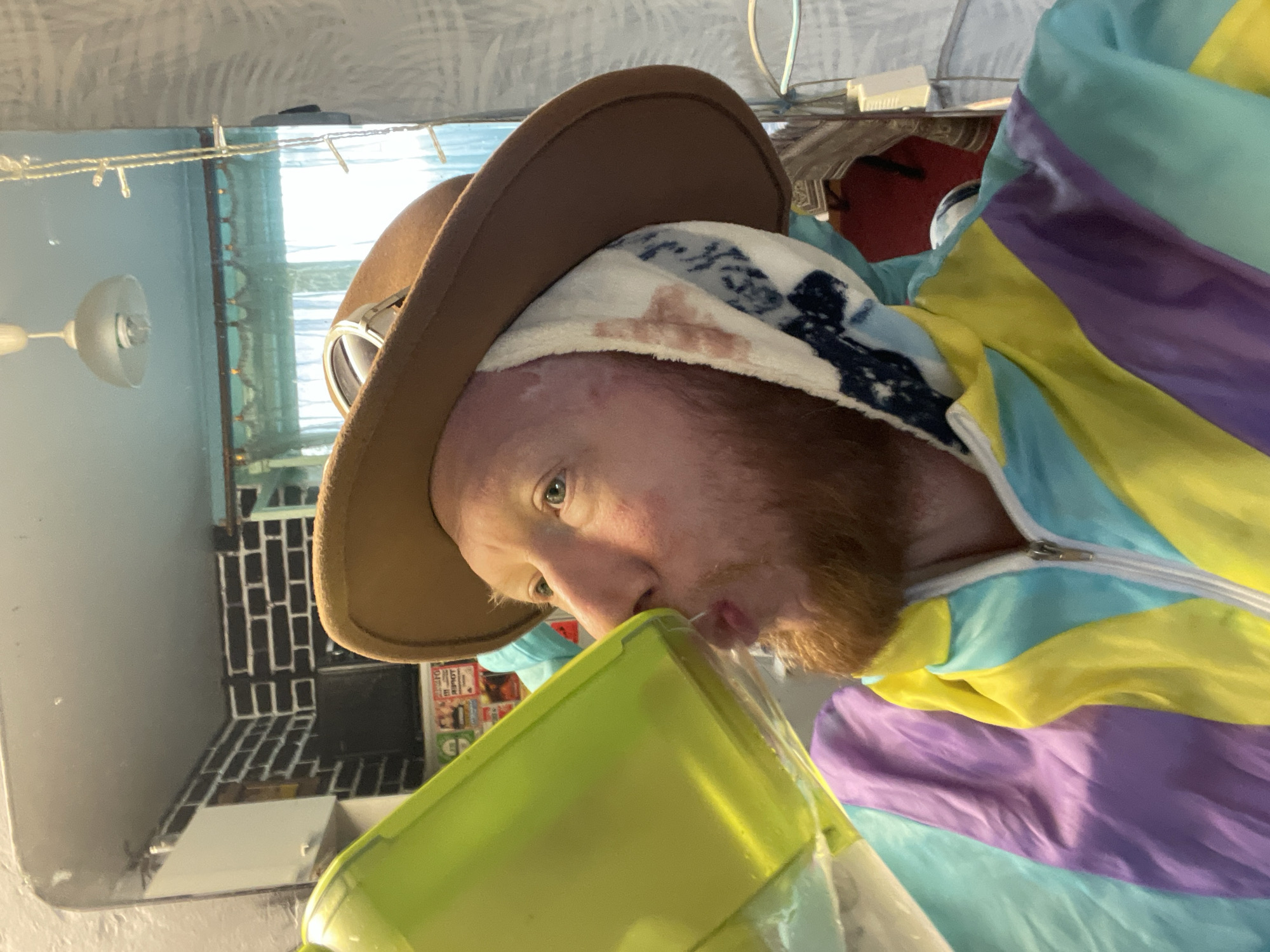 A man with red hair and a beard, wearing a brown fedora and goggles, drinks from a neon green jug. He is also wearing a colorful tracksuit with purple, blue, and yellow panels. A towel or cloth with a blue and white pattern is wrapped around his head. The background appears to be an indoor space with a mirror reflecting a room with a brick-patterned wall, a light fixture, and curtains. The location is the city of Saint Petersburg, Russia.