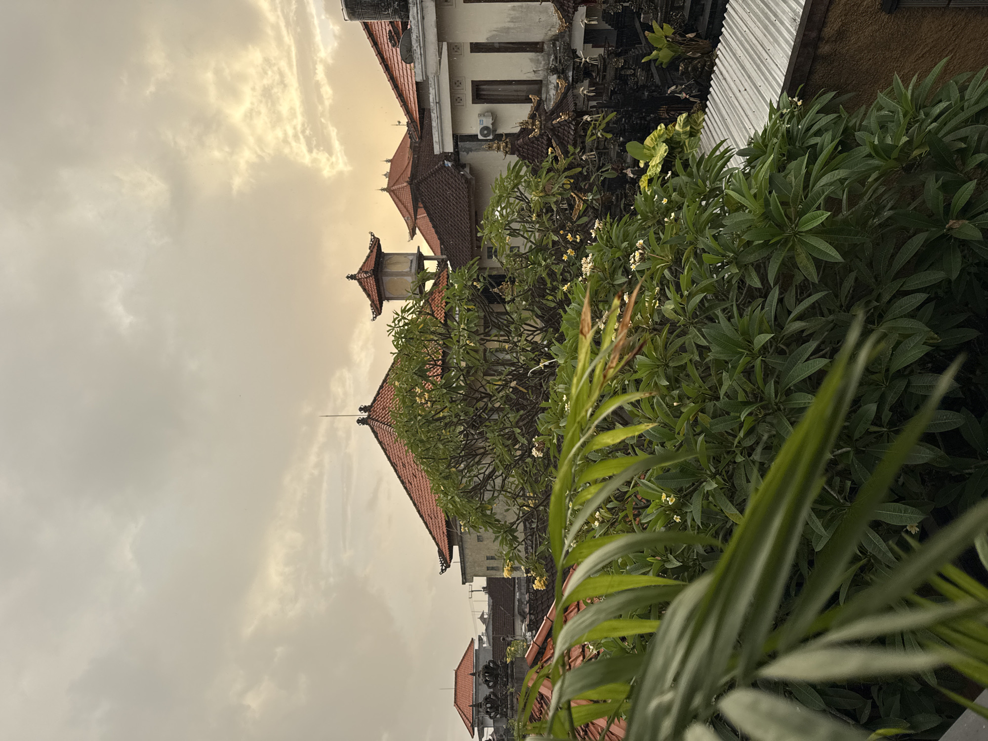 The image shows a view in Kuta, Indonesia. The foreground is dominated by lush green foliage, possibly from a garden or balcony. Behind the foliage, traditional Balinese buildings with distinctive red-tiled roofs are visible. The architecture includes decorative elements and intricate details. The sky is overcast with diffused light, suggesting a cloudy day or early morning. The buildings appear to be residential or small hotels.