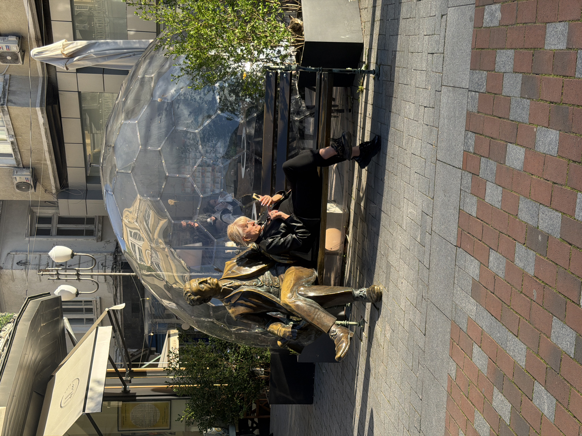The image captures an outdoor urban scene, likely a pedestrian street or public square, bathed in bright sunlight that casts distinct shadows. The central focus is a wooden bench occupied by a woman and a bronze statue, positioned next to a large, clear geodesic dome structure.
A woman with short blond hair, wearing a black jacket, dark trousers, and black boots, is seated on the bench, engrossed in her mobile phone. To her left, a bronze statue of a man, depicted in a suit and seated in a relaxed posture, shares the bench, looking forward. This statue appears to represent a notable figure, adding a historical or cultural element to the modern setting.
Behind the bench and extending to the left is a striking, transparent geodesic dome, constructed from numerous hexagonal panels. Its reflective surface mirrors the surrounding buildings and the sky, suggesting it might be part of a cafe or restaurant seating area. Further in the background, multi-story urban buildings are visible, some featuring windows and air conditioning units. Green foliage from trees or bushes adds a touch of nature to the environment.
The ground is paved with two different patterns: a section of light gray rectangular paving stones and another section of red and gray brickwork arranged in a patterned design. While an awning with an unclear logo is visible on a building to the left, and a small yellow sign with unreadable text is near the statue, no specific legible text provides explicit context. The scene evokes a vibrant public space in a city like Varna, Bulgaria, where people can relax and enjoy the urban atmosphere.