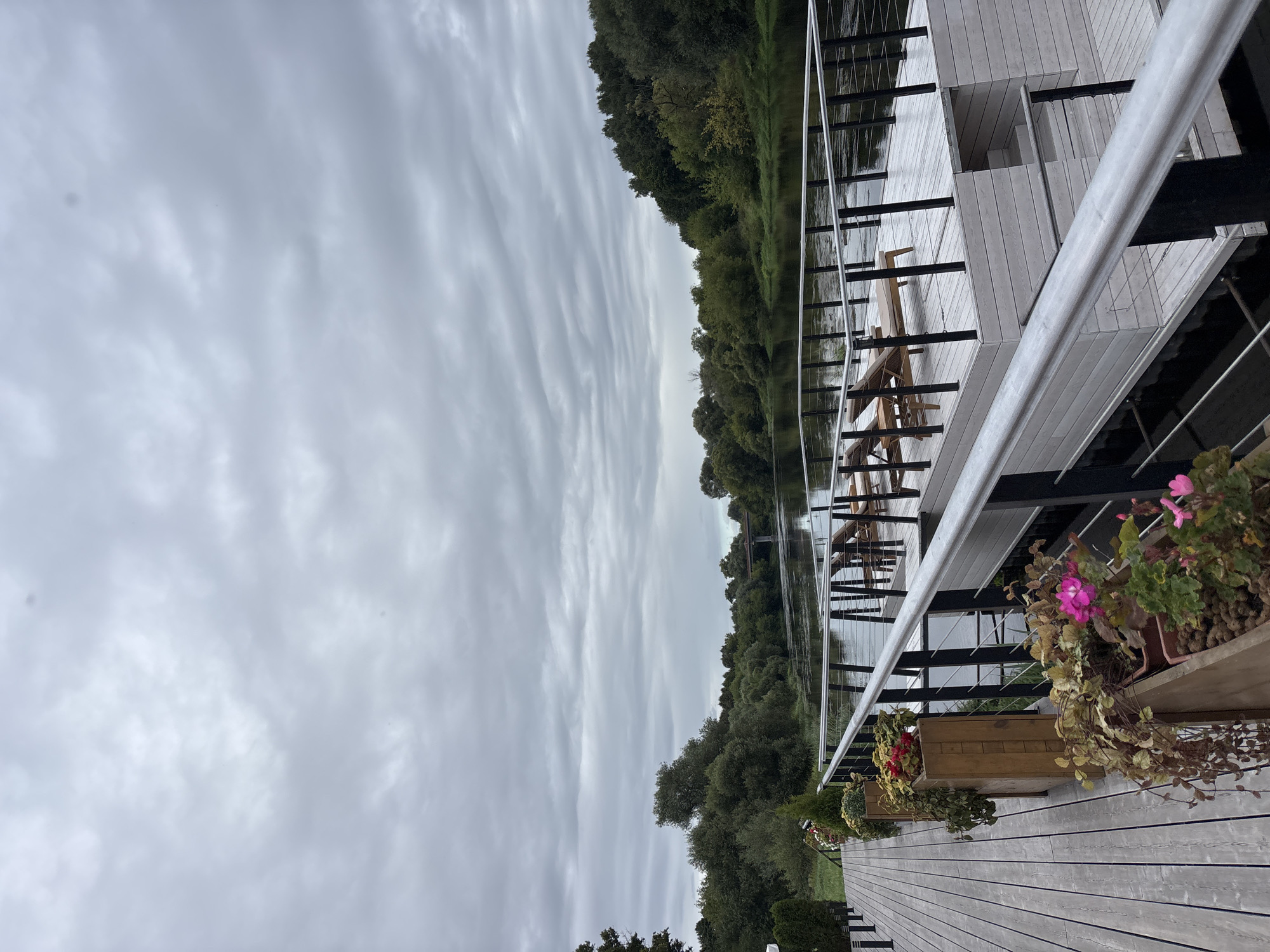 The image depicts a wooden deck on the bank of a river. The deck has planters with flowers and foliage, as well as a railing. In the background, there is a river with a bridge visible in the distance. The opposite bank is lined with trees. The sky is overcast. This scene is located in the city of Zvenigorod, Russia, specifically on the Moskva River near the Savvino-Storozhevsky Monastery.
