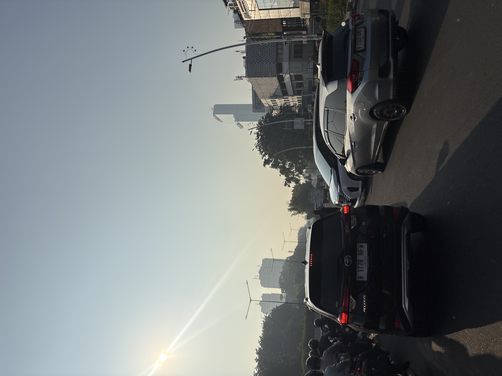 In the city of West Jakarta, Indonesia, a traffic jam is captured on a hazy day. The sun shines brightly from the upper left of the frame, casting long shadows. In the foreground, a black Toyota Rush is stopped, its license plate reading "B 1778 HFW." To its right, a silver Honda Mobilio is also at a standstill, with its license plate partially visible as "B 3141 DR." Several other vehicles, including motorcycles, are present in the congested roadway. In the background, tall buildings and trees are silhouetted against the bright sky. Streetlights are visible along the road, some with decorative elements. The overall atmosphere suggests a typical urban commute in Jakarta during a period of high traffic density.
