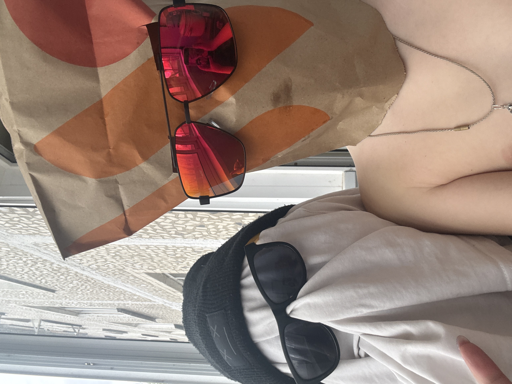 The image, taken in a vertical orientation, captures a close-up, first-person perspective of a person relaxing or reclining. Prominently featured is a crumpled brown paper bag, characteristic of fast food packaging, adorned with a large, abstract logo composed of overlapping orange and reddish-orange arcs and circles, strongly reminiscent of the Taco Bell brand logo.
Resting on this bag is a pair of sunglasses with bright red, mirrored lenses and black frames. Reflections within these lenses reveal an indoor environment, possibly a vehicle interior or a room. Partially visible to the right is the upper chest and shoulder of a person with fair skin, wearing a light-colored top. A delicate, silver-toned chain necklace with a small, metallic pendant is worn around their neck.
In the lower left, a black knitted beanie or cap is visible, featuring a patch with two distinctive white 'X' shapes. Another pair of black-framed sunglasses with dark lenses rests on this beanie and a section of light-colored fabric, likely part of the person's clothing or a blanket. A finger, possibly the pinky or ring finger of the person, is visible near the bottom right edge of the frame.
The background shows a light-colored, patterned surface, which could be a ceiling or an awning with a geometric, tessellated design. Below this, a window or partition with a light-colored frame is present. The bright lighting suggests it is daytime. The overall scene conveys a casual, relaxed moment, perhaps after a meal, occurring within an indoor or vehicle setting in Dmitrov, Russia.