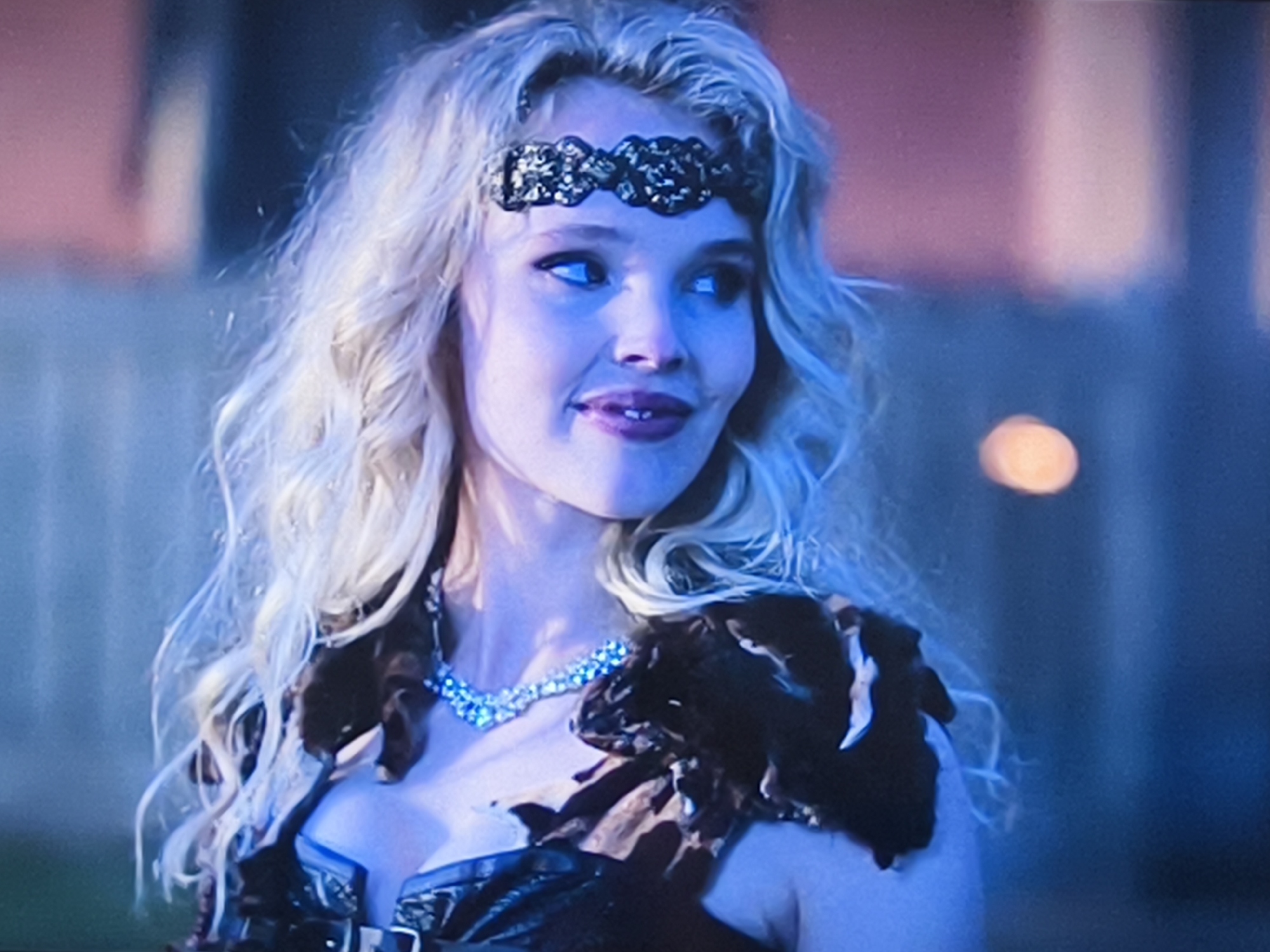 A young woman with blonde wavy hair and a black sequined headband is looking slightly to the right. She is wearing a low-cut brown leather garment with fur on the shoulders and a diamond necklace. The background is blurred, suggesting an outdoor environment at night with a light source in the distance. There is no information to suggest this image was taken in Petah Tikva, Israel.