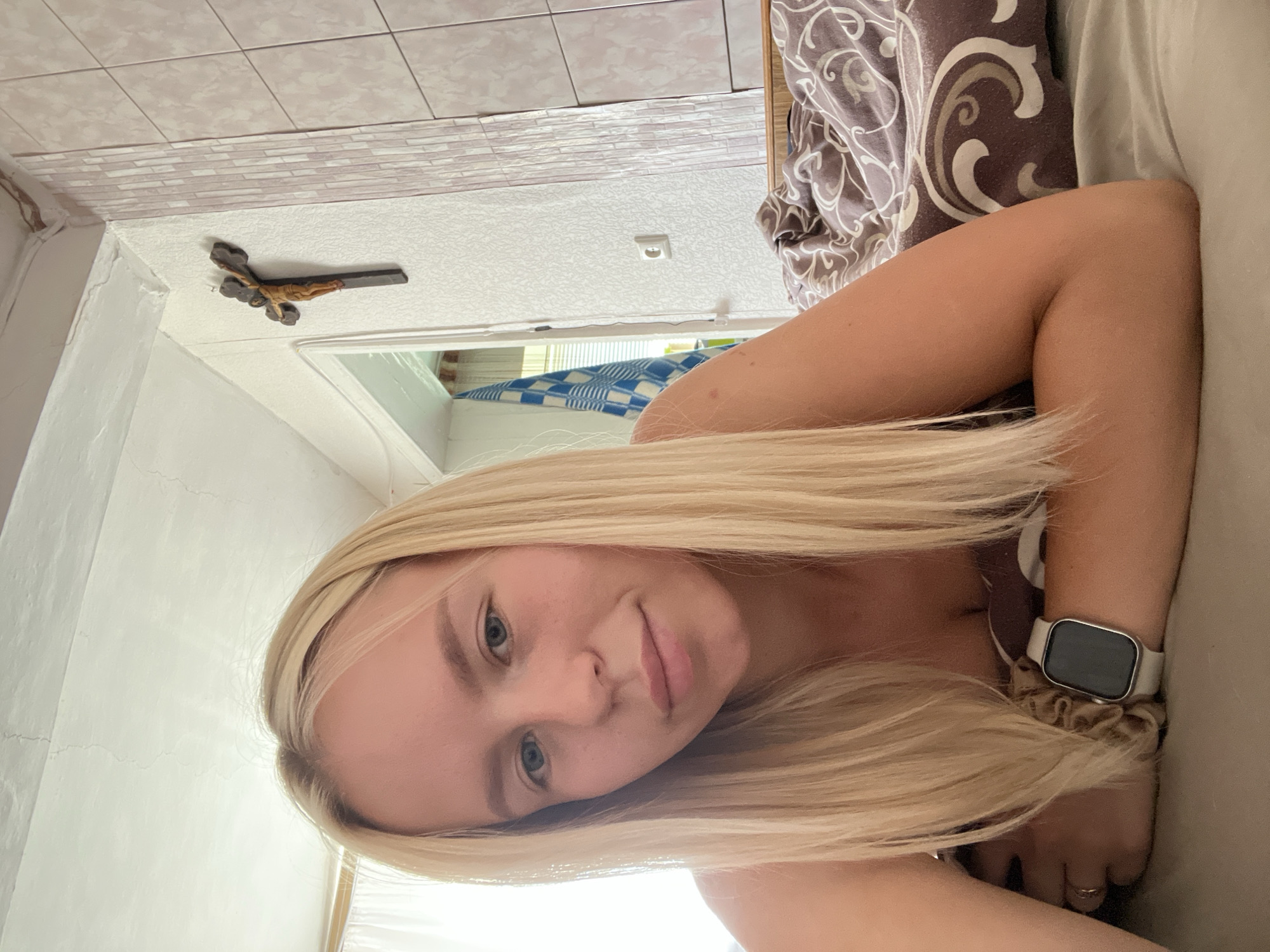 The media file is an image of a young woman with blonde hair lying down. She is wearing a smartwatch on her left wrist and a ring on her right ring finger. Her head is tilted and she is looking towards the camera. In the background, there is a wall with a crucifix mounted on it. There is also a patterned fabric and a checkered blanket visible. The lighting suggests it is daytime, and the setting appears to be indoors, possibly a bedroom. The woman's expression is neutral or slightly smiling.