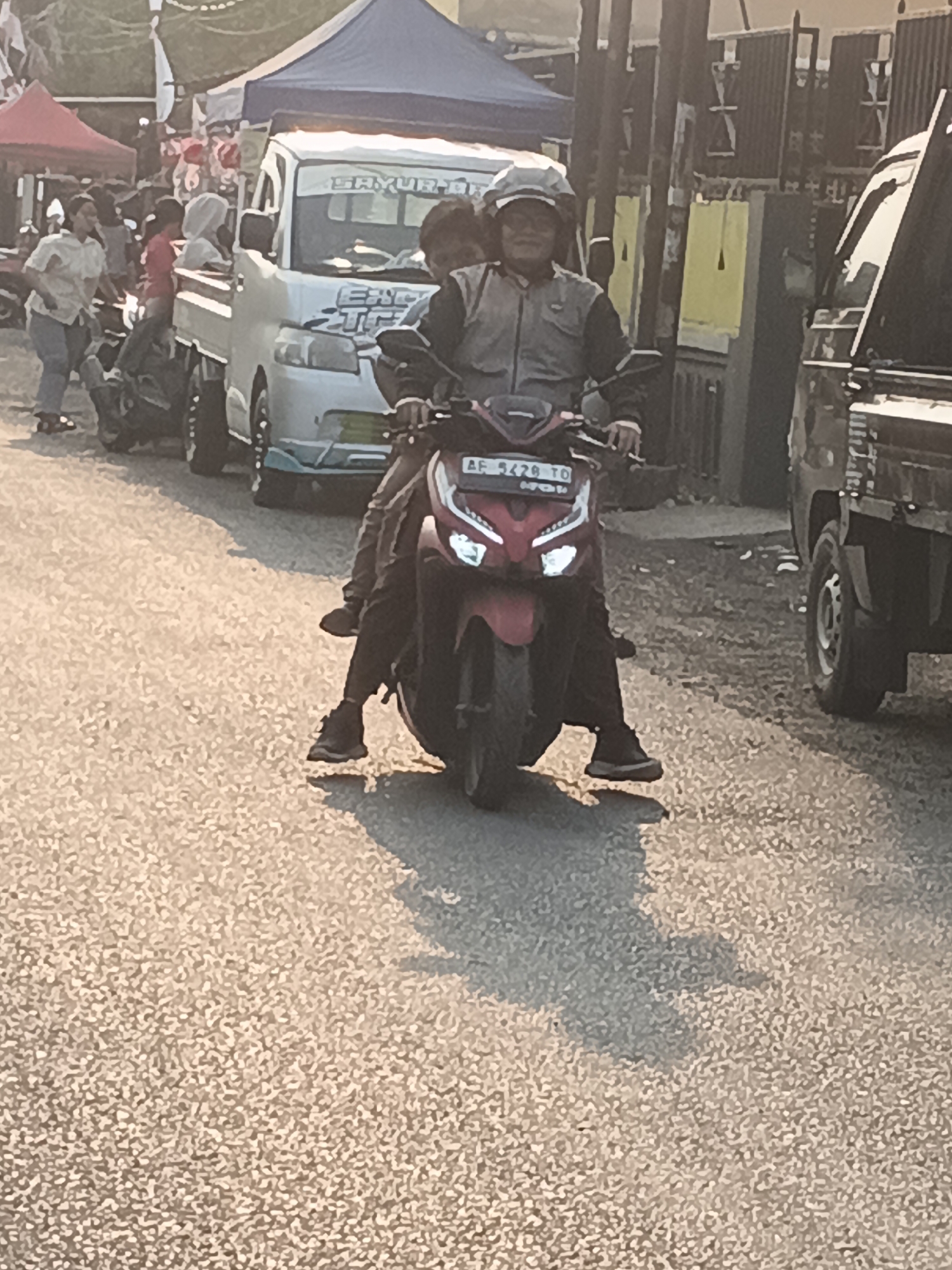 The image captures a street scene in Bogem Pinggir, Indonesia, during daylight. In the foreground, a person rides a red scooter, facing left, with their left leg extended forward. The scooter has a license plate with "AF 5429 TO" visible. Behind the scooter, a white pickup truck is parked, with a blue canopy overhead and text reading "SAYUR" on its windshield. Several other vehicles, including a dark-colored pickup truck, are also present on the street. The background shows tents and buildings, suggesting a market or event. The ground is paved with asphalt. People are visible in the background, some standing and others possibly on motorcycles. The lighting indicates it is daytime, with sunlight casting shadows on the ground.